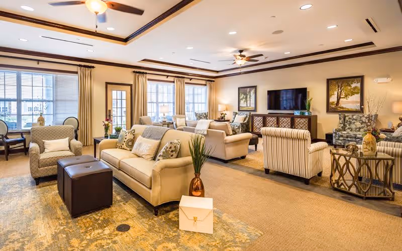 A spacious and well-lit living room with beige sofas, patterned armchairs, a large area rug, and a flat-screen TV mounted on the wall. The room features large windows with curtains, ceiling fans, framed artwork, and decorative plants on side tables.