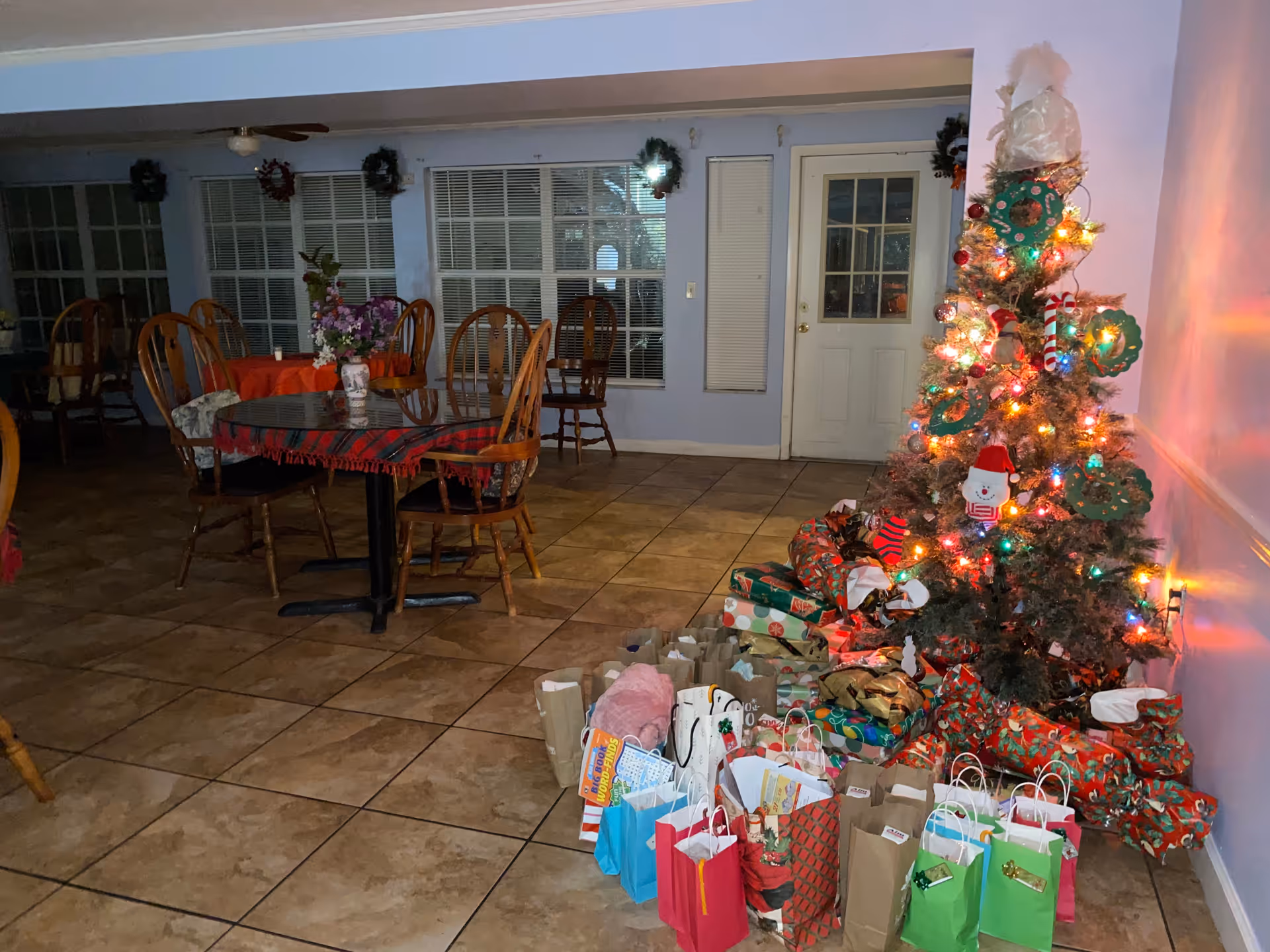 A decorated Christmas tree with colorful lights and ornaments stands in the corner of a room. Numerous wrapped gifts and gift bags are placed on the floor around the tree. The room has tiled flooring, several wooden chairs, and tables with tablecloths. The walls are light blue with windows and a white door in the background.