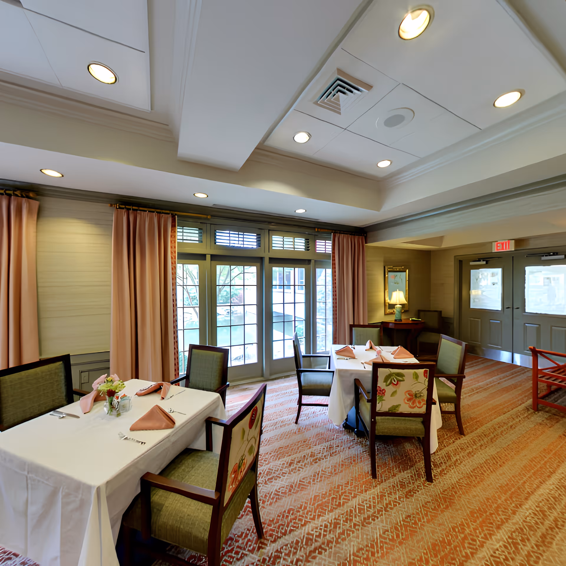 A dining room with two tables covered in white tablecloths, each set with folded pink napkins, cutlery, and small flower arrangements. The room has large windows with pink curtains, a patterned carpet, and recessed ceiling lights. There is a door with an exit sign above it and a small table with a lamp in the corner.