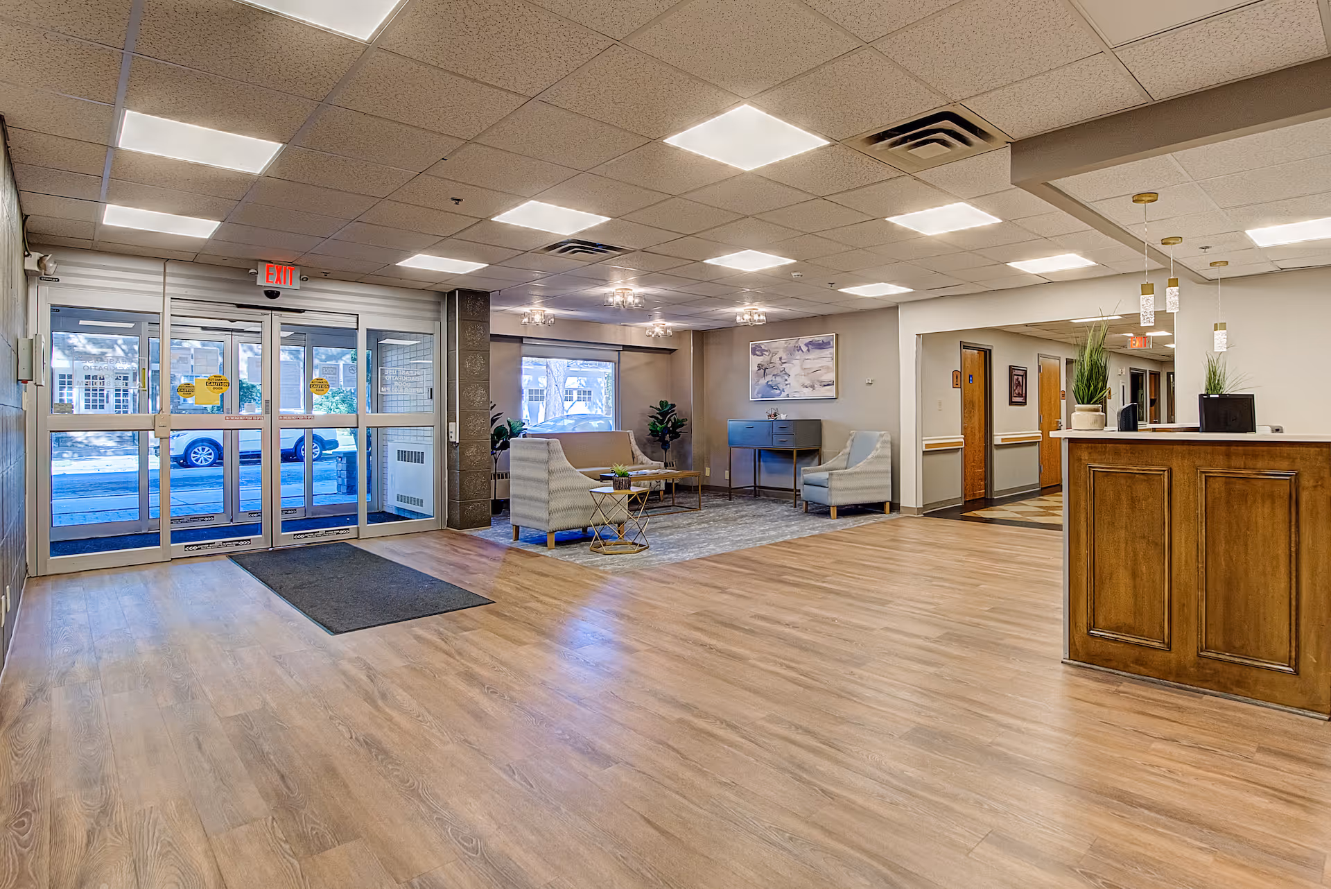 Spacious and well-lit senior living facility lobby with wooden flooring, a seating area with chairs and a coffee table, a reception desk with pendant lights, and large glass entrance doors showing a view of the outside street.