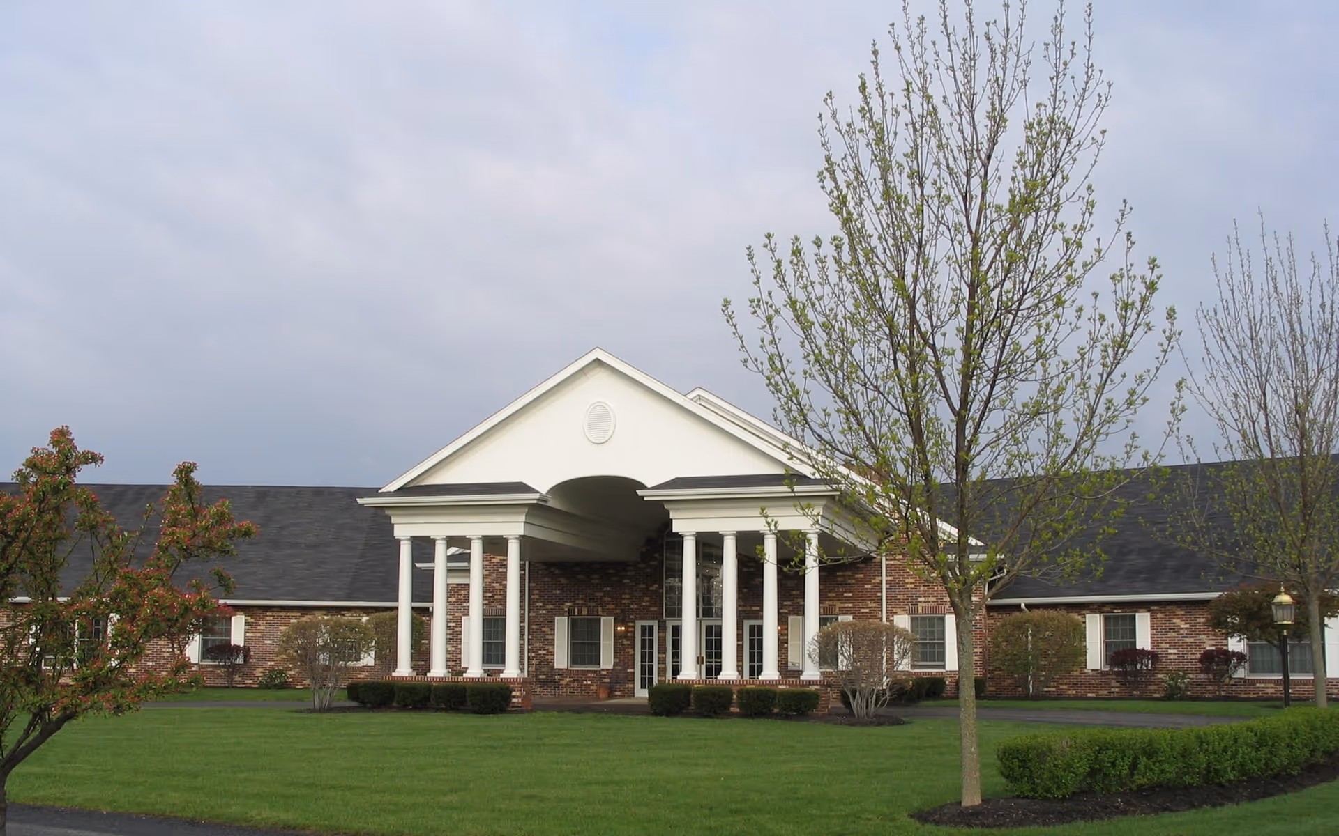 Front exterior view of a single-story brick building with white columns supporting a covered entrance, surrounded by a well-maintained lawn and young trees under a cloudy sky.