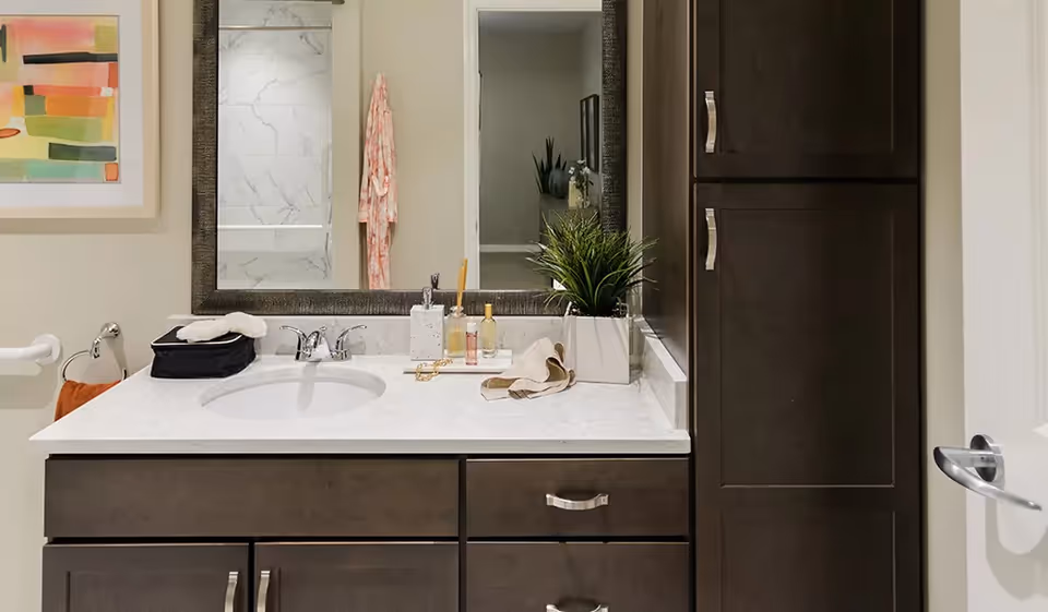 A bathroom vanity with a white marble countertop, an oval sink, and silver faucet. On the countertop are a black toiletry bag, a soap dispenser, a small tray with bottles, a beige cloth, and a potted green plant. Above the vanity is a large rectangular mirror reflecting a shower with marble tiles and a pink robe hanging on a hook. To the right of the vanity are dark wood cabinets with silver handles. A colorful abstract painting is partially visible on the left wall.