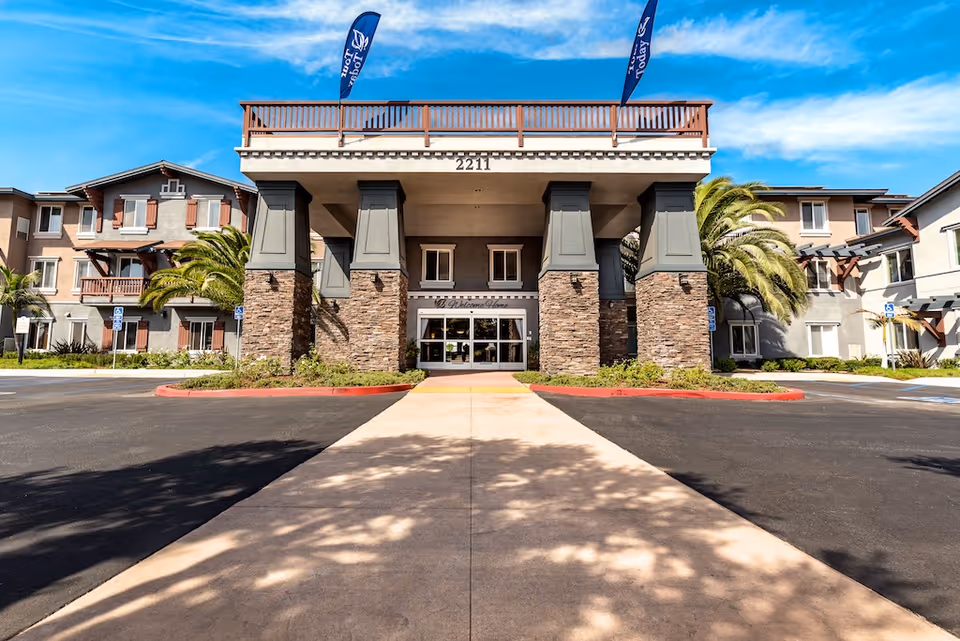 Front entrance of a multi-story senior living building with a covered porte-cochere supported by stone columns and flags overhead.