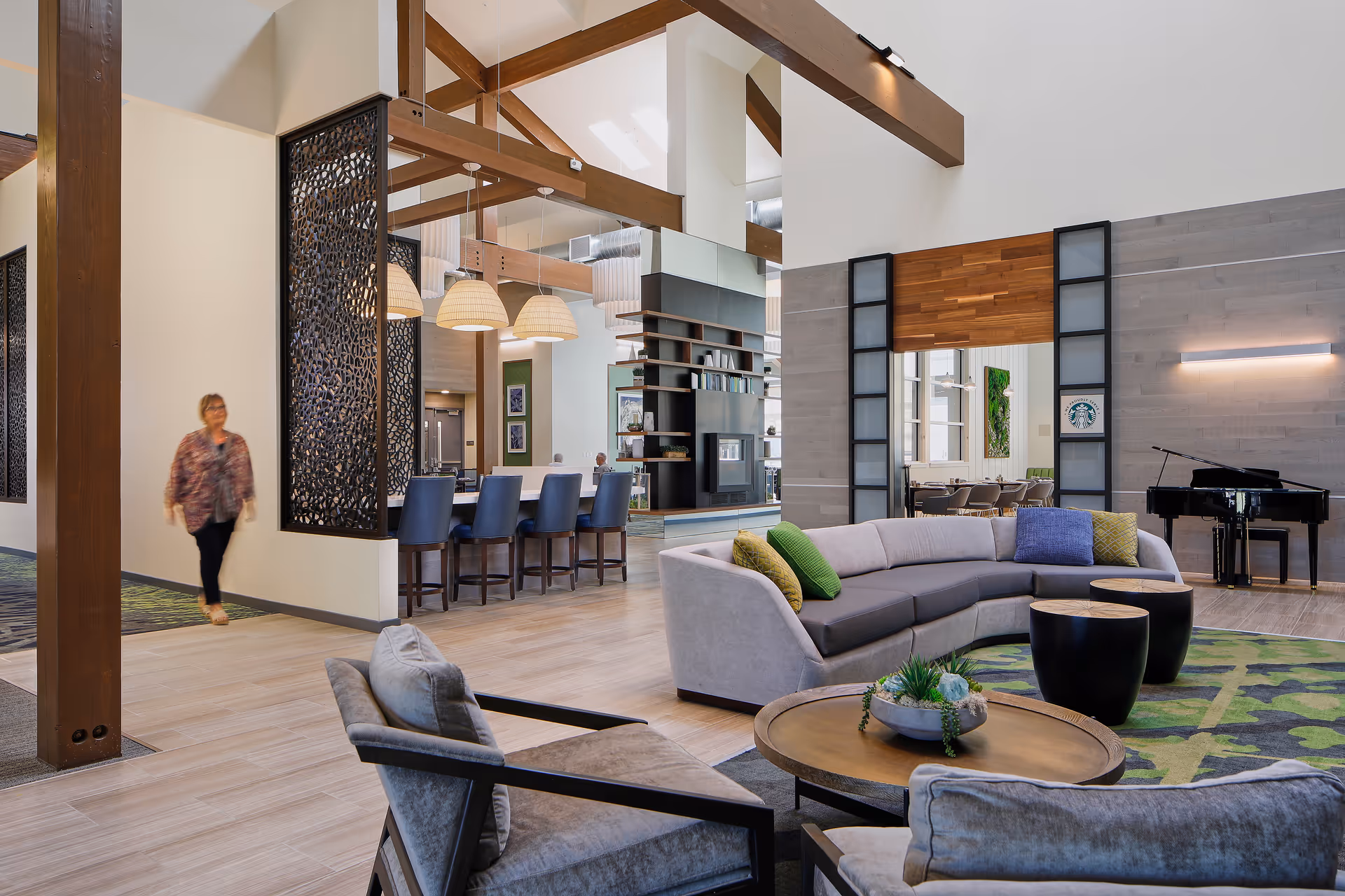 A spacious and modern living room area in a senior living facility with high ceilings and exposed wooden beams. The room features a curved gray sofa with colorful pillows, two round black coffee tables, and a round wooden table with a plant centerpiece. In the background, there is a bar counter with blue chairs and hanging pendant lights. A grand piano is visible near a wall with a light fixture, and a woman is walking through the space. The area is well-lit with natural light coming through large windows.