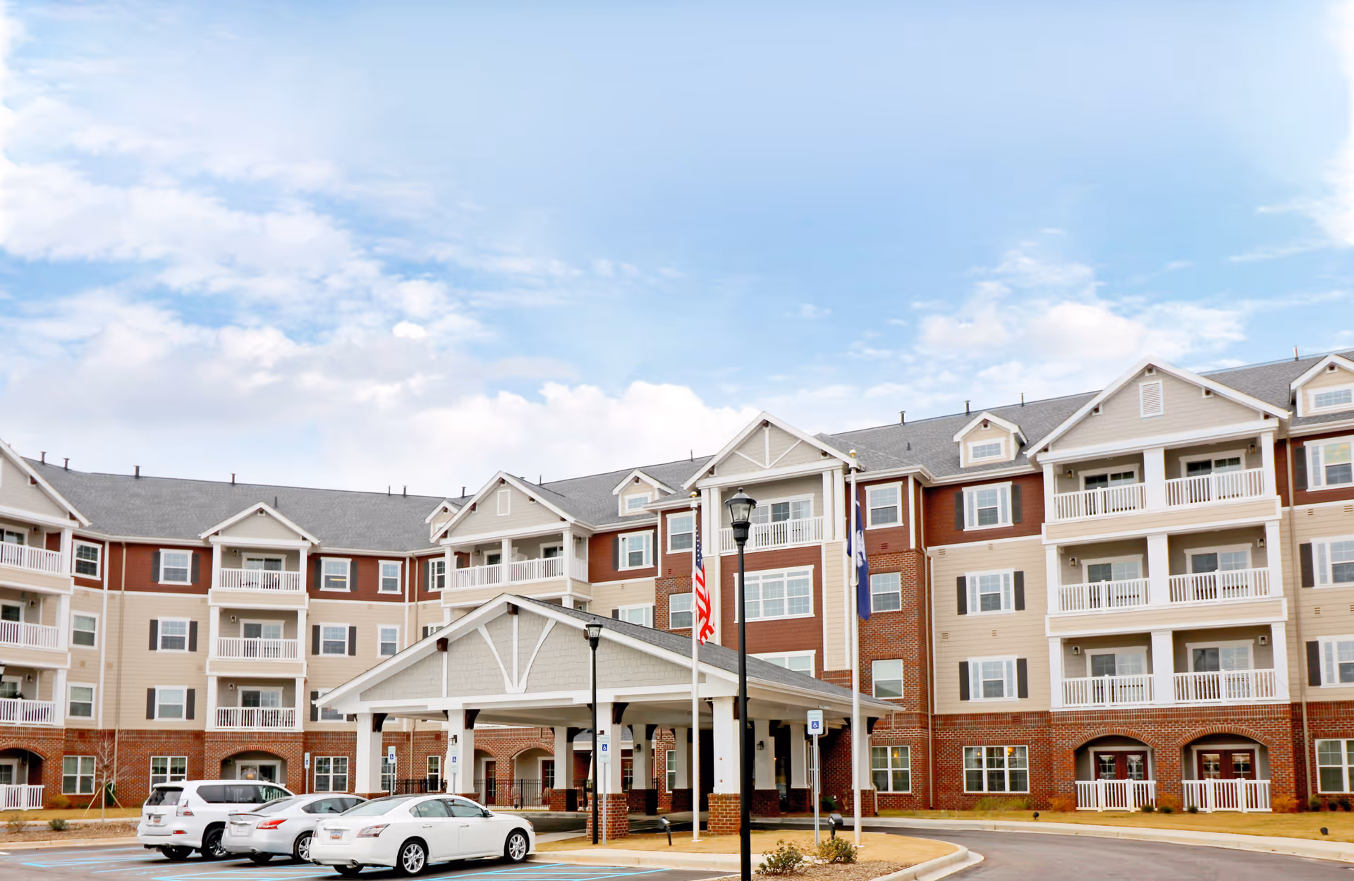 Exterior view of a multi-story senior living facility named Harmony at Five Forks with a covered entrance, several parked cars, and flags displayed near the entrance under a partly cloudy sky.