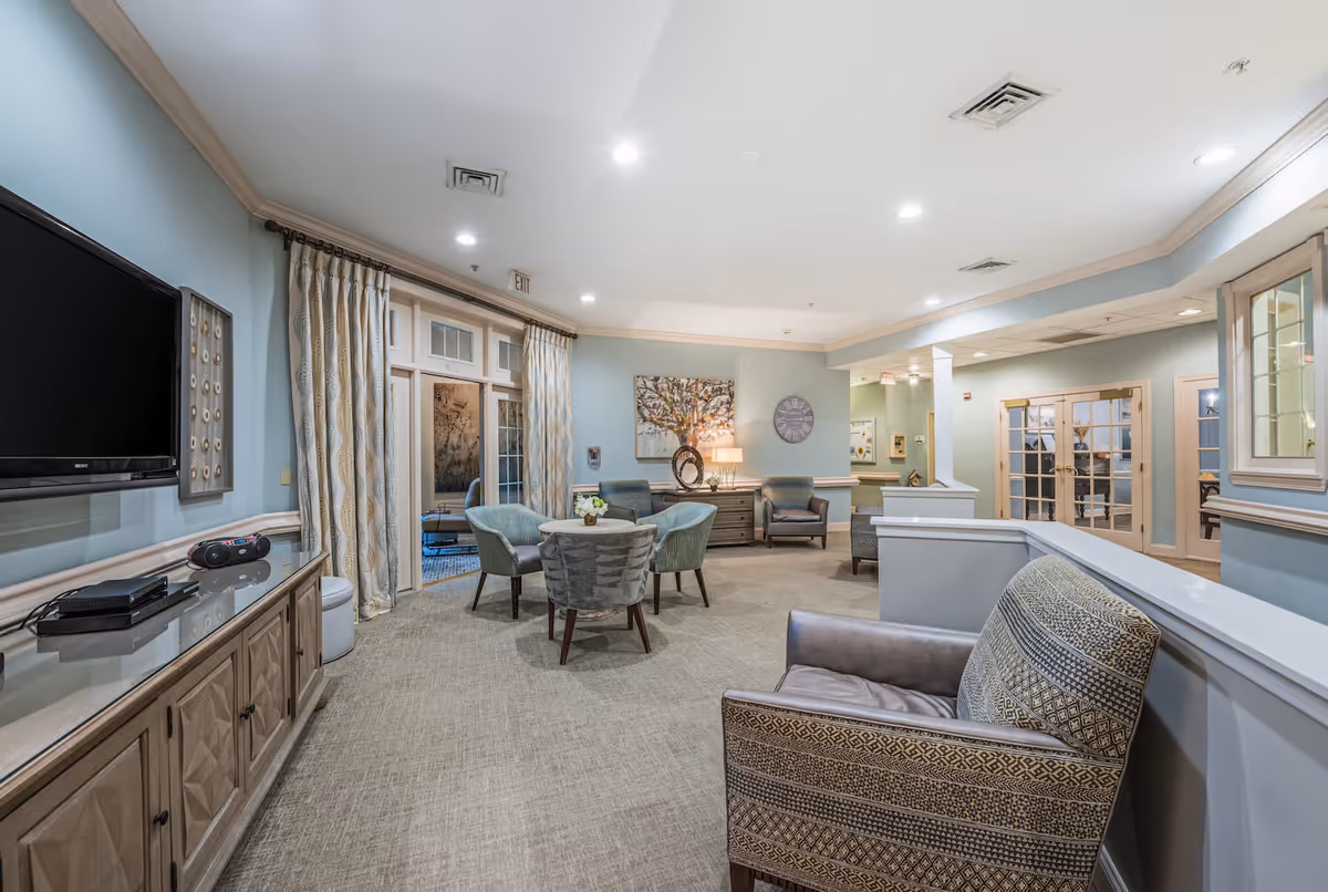 Well-lit senior living lounge with armchairs and a round table, a wall-mounted TV, and French doors.