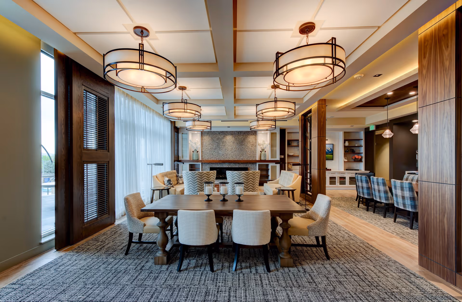 A spacious and well-lit common area featuring a large wooden dining table surrounded by upholstered chairs. The room has modern circular pendant lights hanging from a coffered ceiling. In the background, there is a cozy seating area with sofas and armchairs arranged around a fireplace with a textured stone wall. Large windows with sheer curtains allow natural light to fill the space. The room has a mix of carpet and wood flooring, with wooden columns and built-in shelving visible.