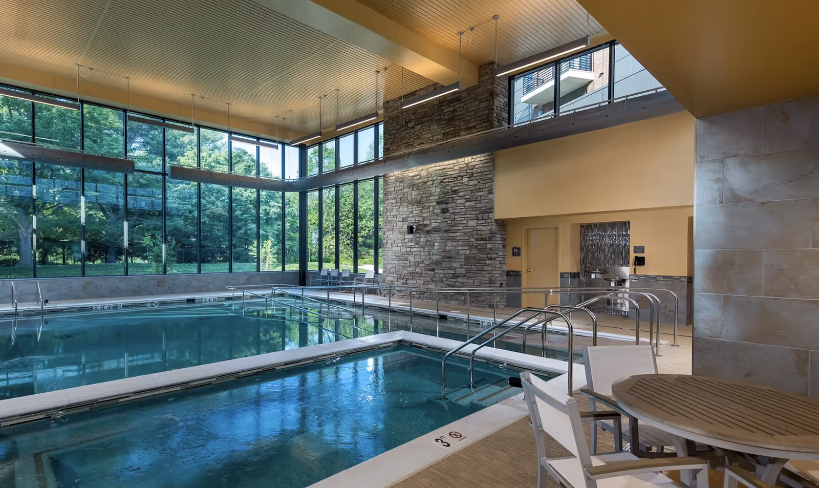 Indoor pool area with large floor-to-ceiling windows showing green trees outside. The pool has metal handrails and a shallow section marked 3 feet deep. There is a round table with chairs in the foreground and a stone accent wall in the background. The ceiling is wooden with hanging lights.