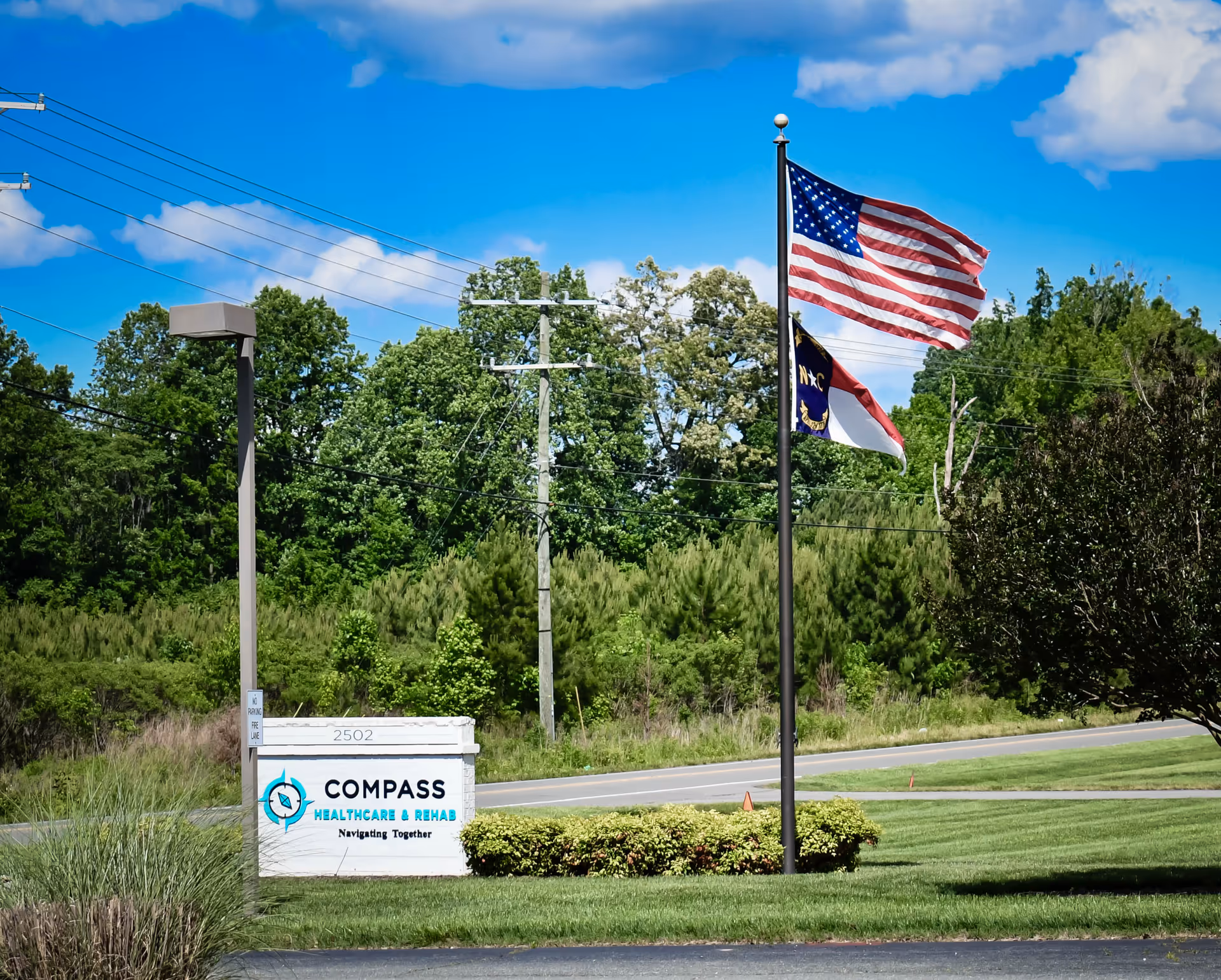 Outdoor view of a sign for Compass Healthcare & Rehab with the tagline Navigating Together, located on a grassy area near a road. Two flagpoles display the American flag and a North Carolina state flag, with trees and a blue sky with clouds in the background.
