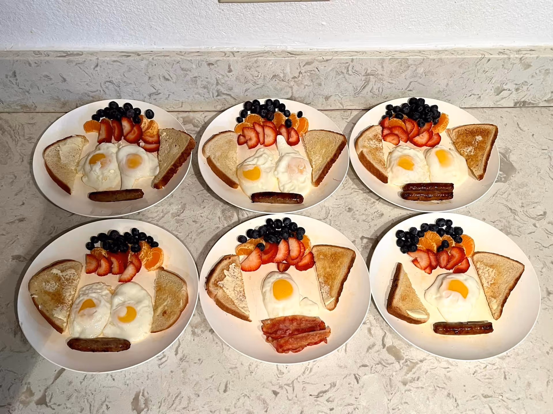 Six plates of breakfast arranged on a marble countertop, each plate containing two slices of toast, two fried eggs, a sausage or bacon strip, and a serving of fresh fruit including blueberries, sliced strawberries, and mandarin orange segments.