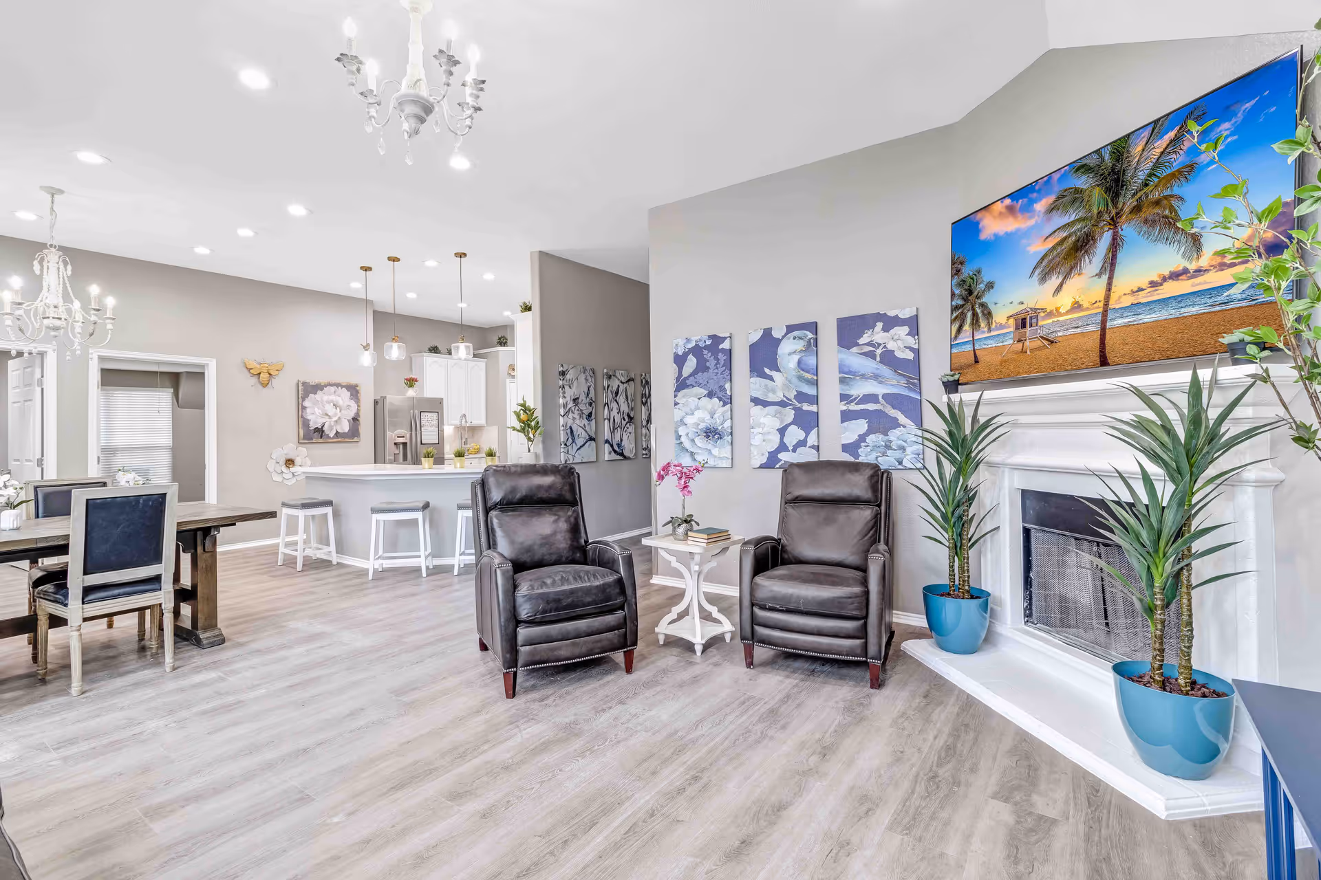 Bright common living area with two leather recliners by a fireplace and wall-mounted TV, with a dining table and kitchen island visible in the background.