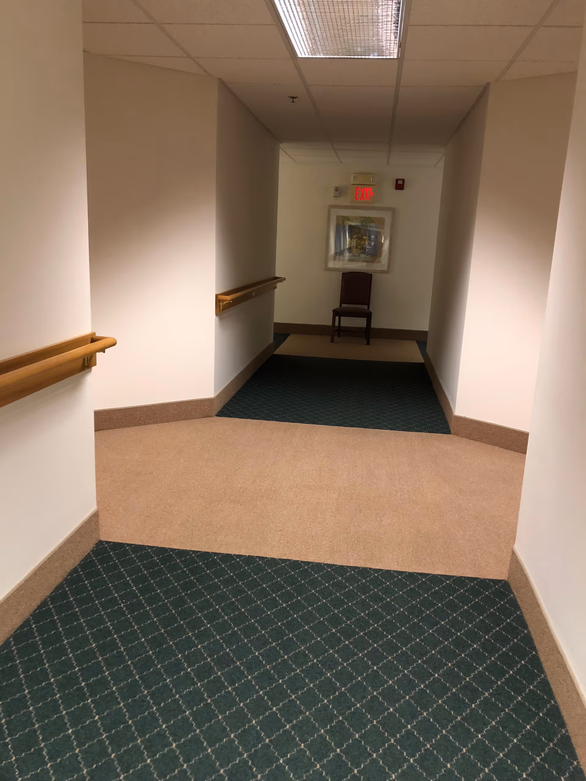 A hallway in a senior living facility with beige and green patterned carpet, white walls, wooden handrails on both sides, a chair at the end of the hallway, a framed picture on the wall, and an illuminated red exit sign above the chair.