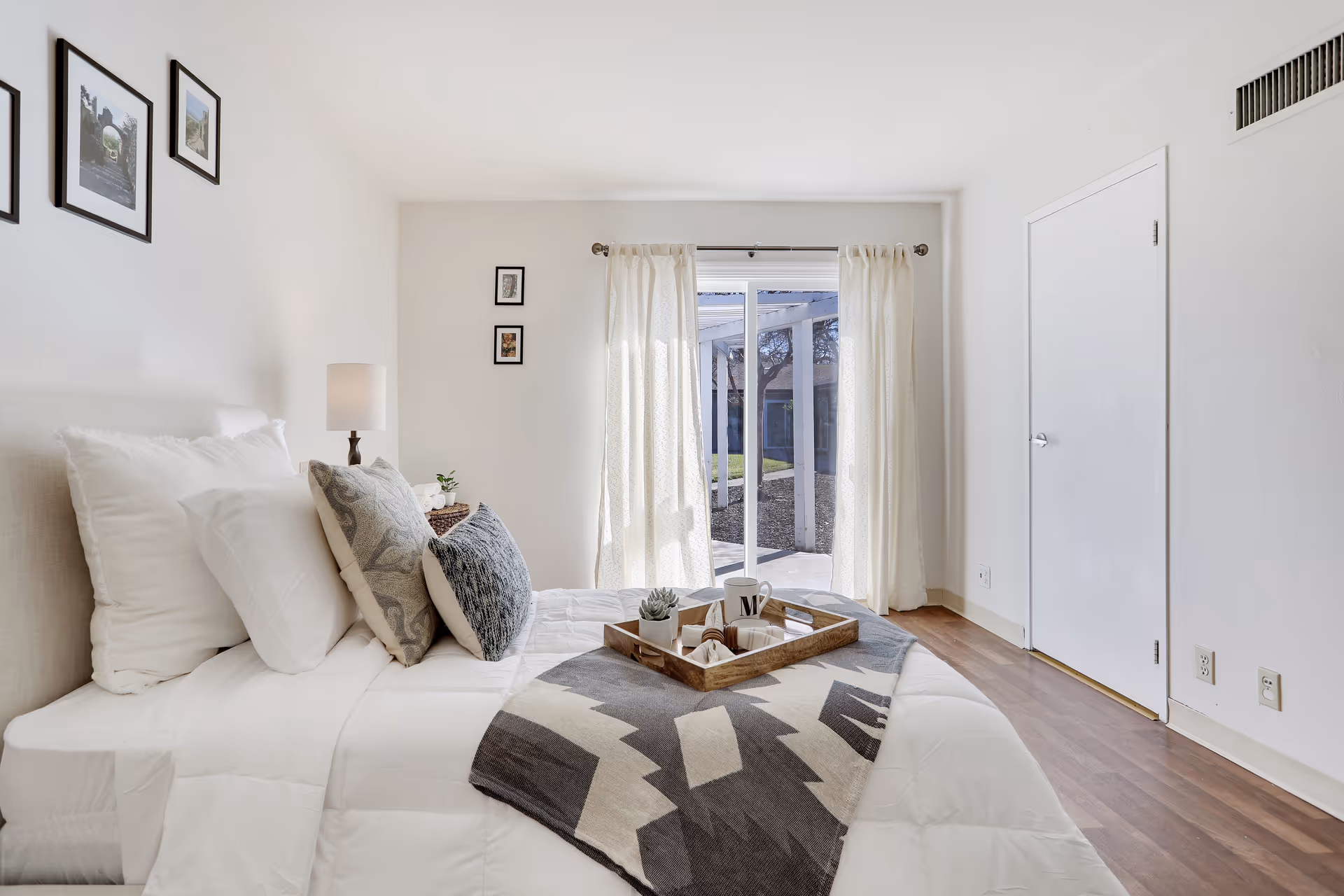 Bright, neutral bedroom with a made bed, decorative pillows and a tray, and a sliding glass door with curtains leading outside.