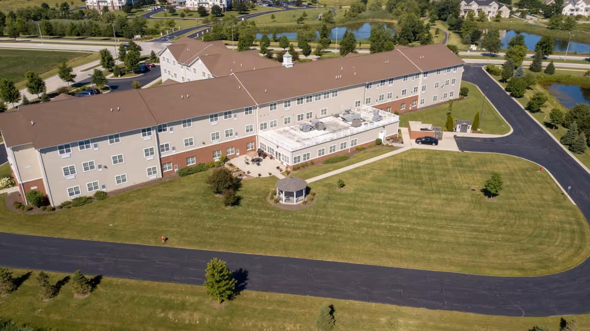 Aerial view of a large senior living facility building with beige and brick exterior, surrounded by well-maintained green lawns, a small gazebo, paved roads, and nearby ponds.