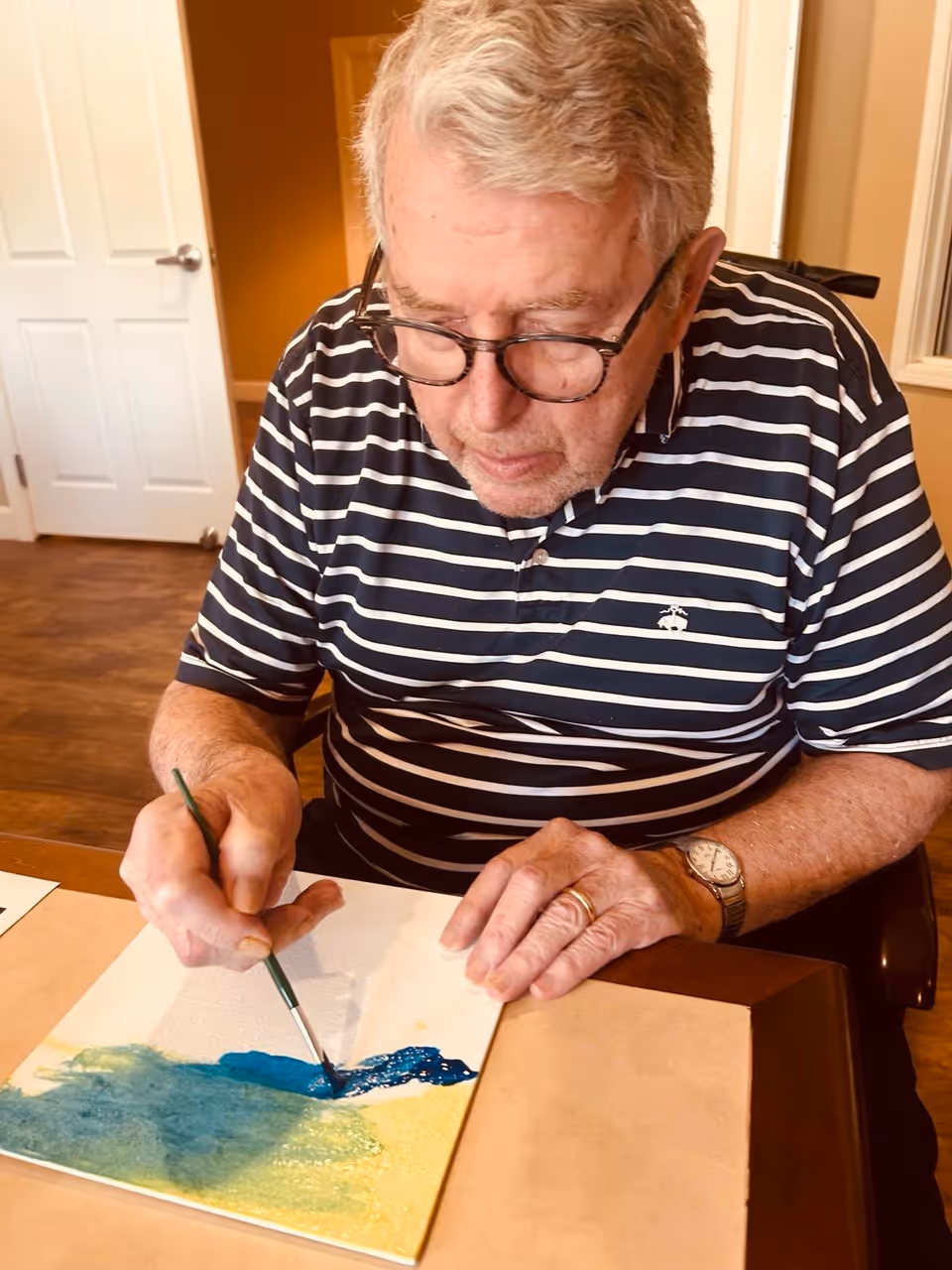 An elderly man wearing glasses and a striped polo shirt is seated at a table, painting on a canvas with blue and green colors. He is focused on his artwork, holding a paintbrush in his right hand and steadying the canvas with his left hand. The room has wooden flooring and a white door in the background.