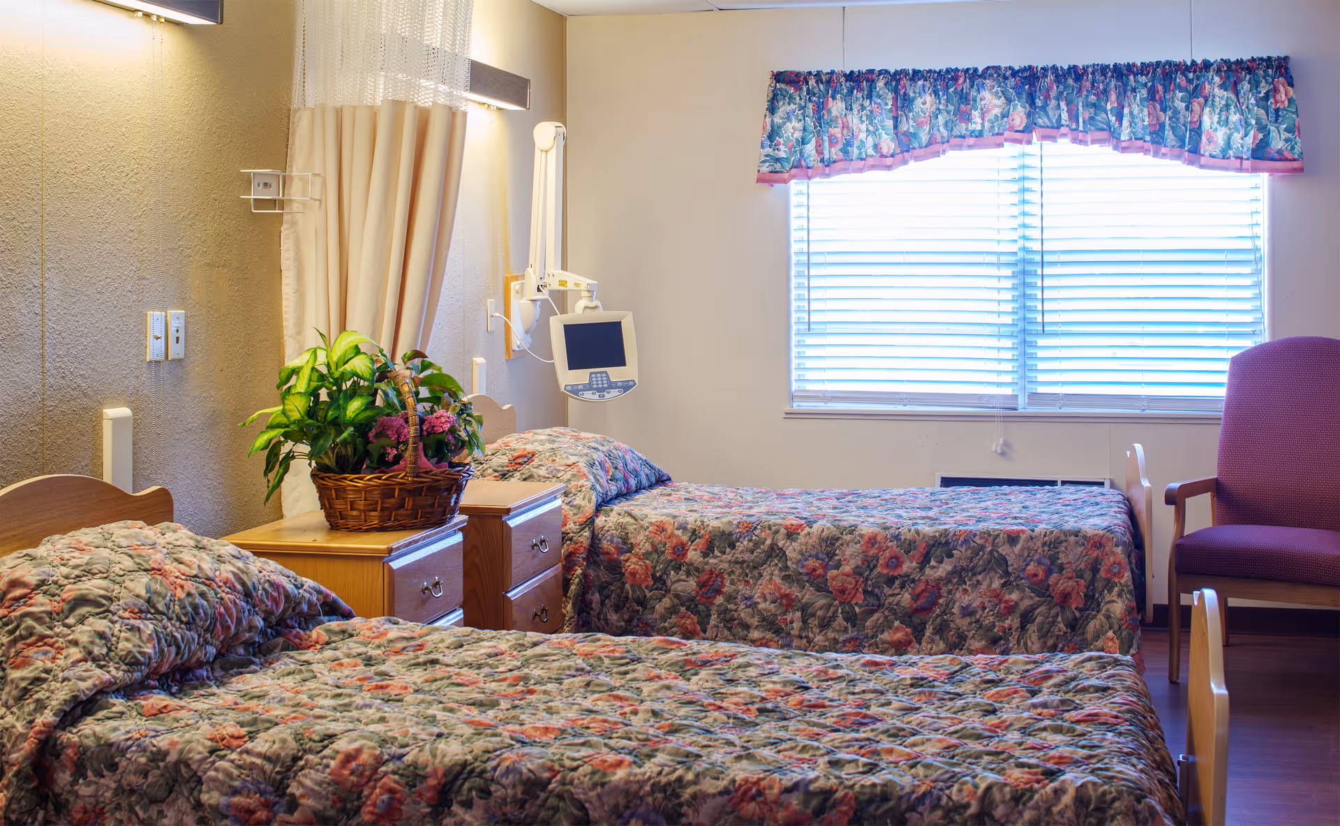A room with two single beds covered in floral bedspreads, a wooden nightstand with a basket of green plants and flowers, a medical monitor mounted on the wall, a window with blinds and floral valance, and a purple armchair.