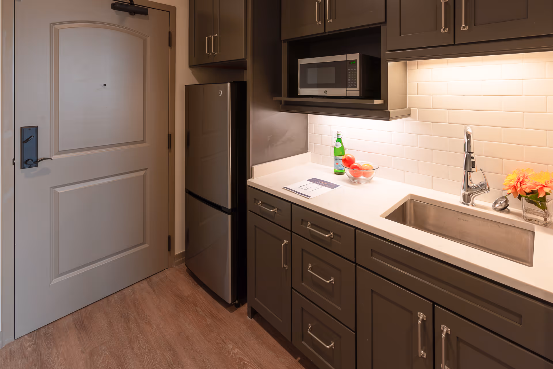 Small kitchenette with gray cabinets, a refrigerator and microwave, a countertop sink, and a closed entry door.