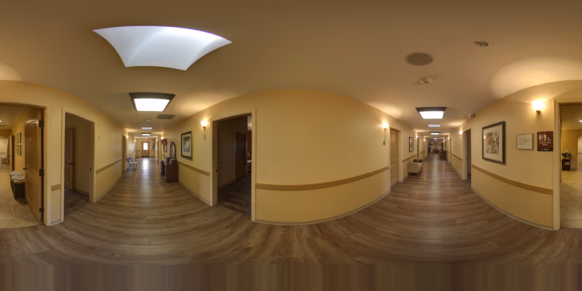 A wide panoramic view of a well-lit hallway in an assisted living facility with wooden flooring, beige walls, and ceiling lights. Several doorways lead to rooms on both sides of the corridor, with framed artwork and signs on the walls. The hallway appears clean and spacious with some chairs and furniture visible in the distance.