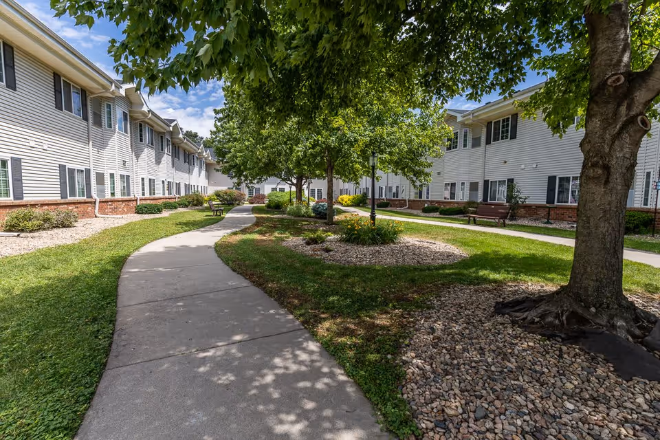 A curved concrete walkway runs through a landscaped outdoor area with green grass, trees, and flower beds. On either side of the walkway are two-story buildings with white siding and multiple windows. Benches are placed along the path under the shade of the trees. The sky is partly cloudy with blue patches visible.