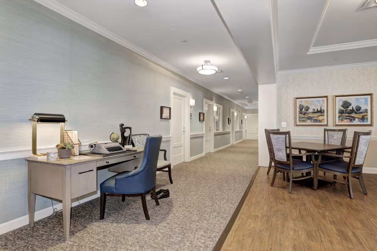 Well-lit senior living common hallway with a desk and chair on the left and a round dining table with chairs and framed artwork on the right.