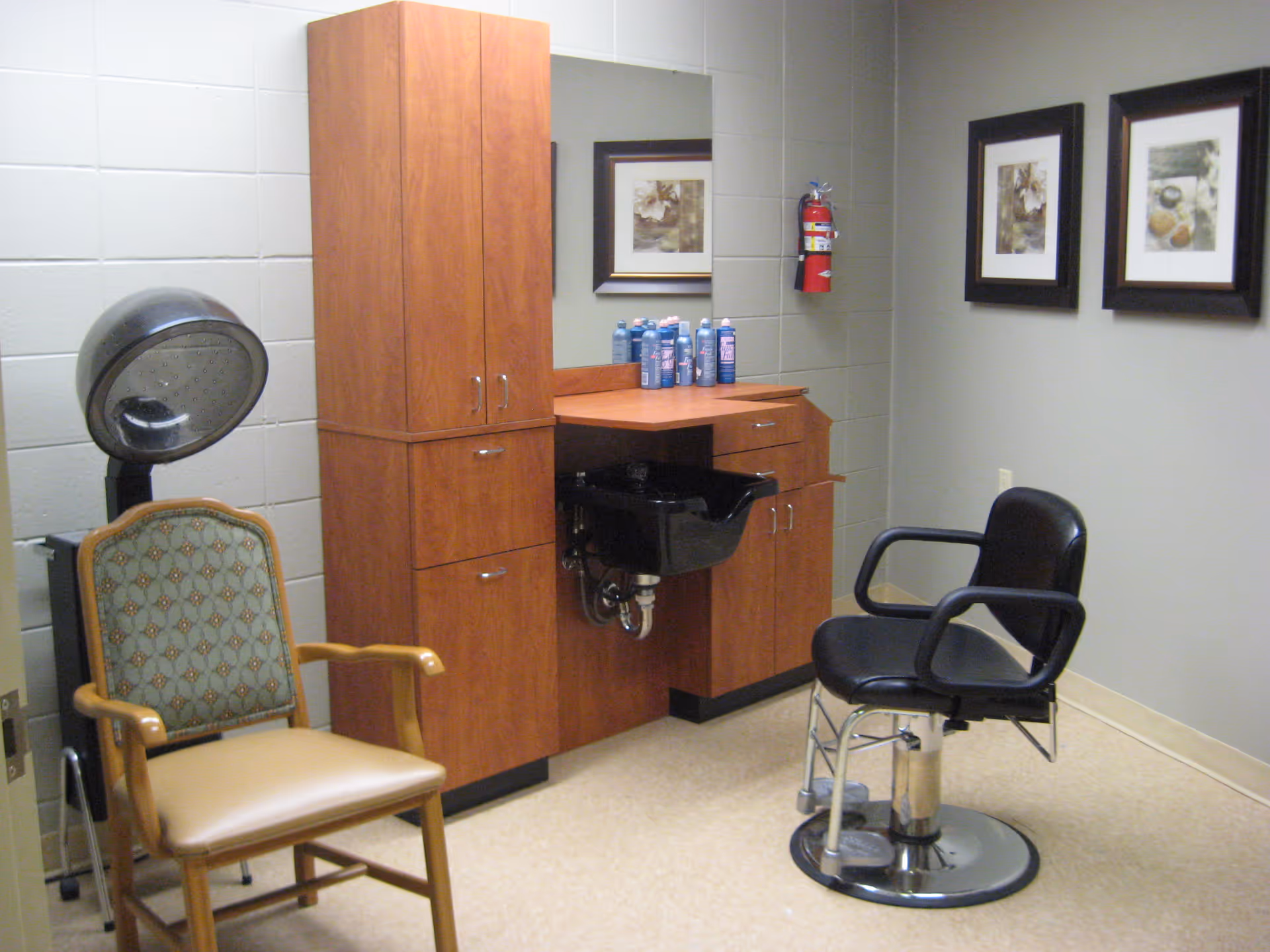 Small salon room with two chairs, a hair-washing sink, wooden cabinets and framed artwork on the wall.