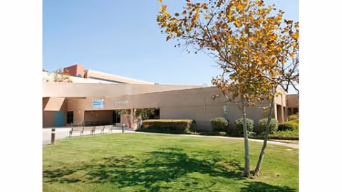 Exterior view of a single-story building with a flat roof, surrounded by green grass, bushes, and a tree with yellow leaves under a clear blue sky.