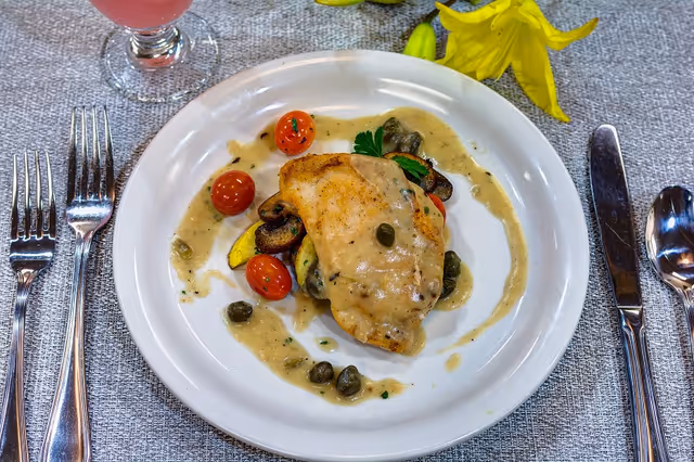 A plated chicken entree with creamy sauce, capers, cherry tomatoes and vegetables on a white plate surrounded by silverware and a yellow flower.