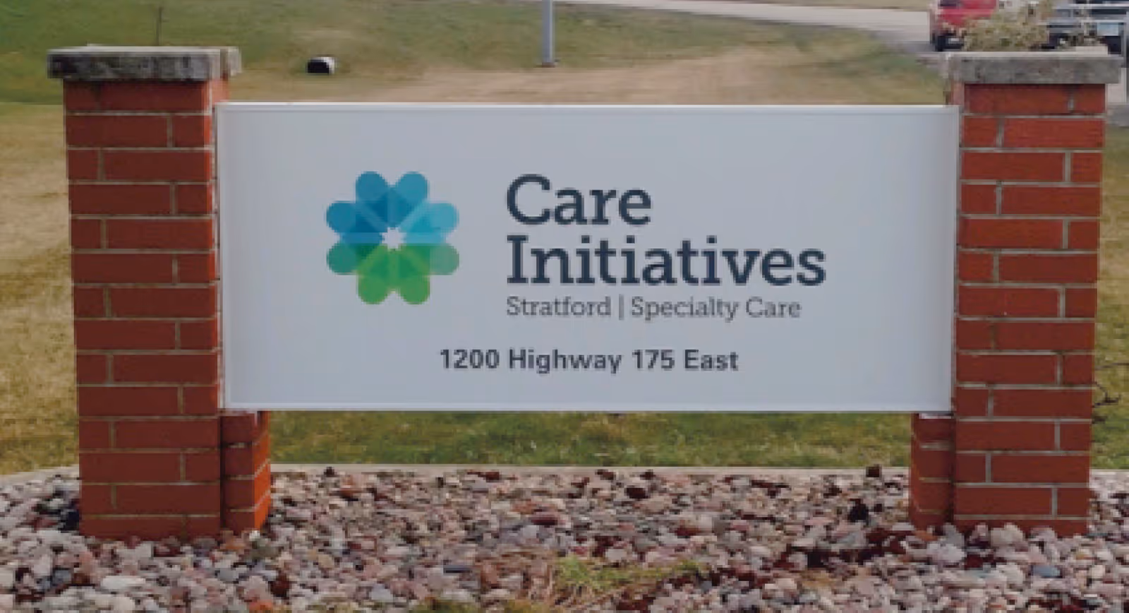 Outdoor sign for Care Initiatives Stratford Specialty Care located at 1200 Highway 175 East, mounted between two brick pillars with grass and a road in the background.