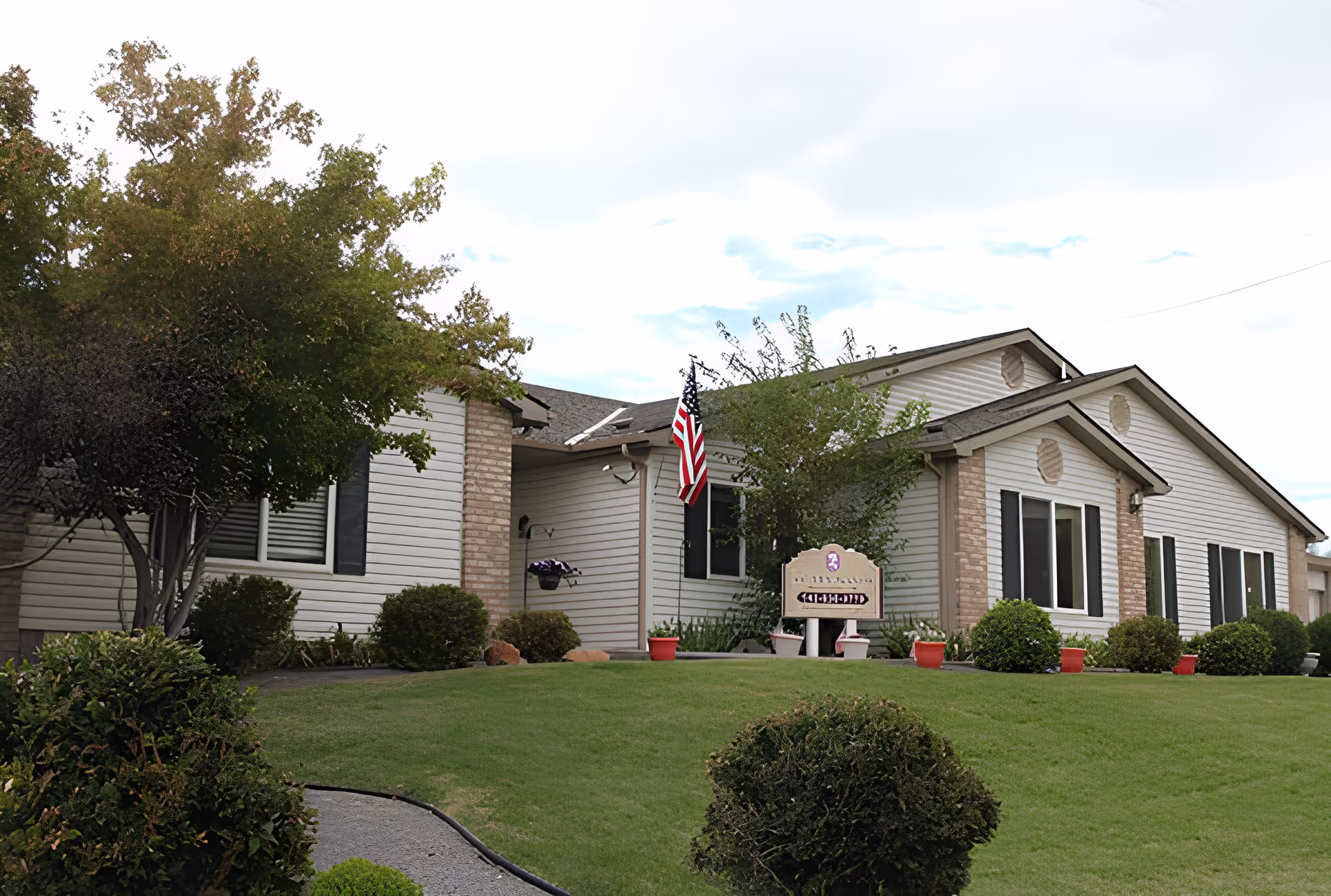 Exterior view of a single-story building with white siding and brick accents, surrounded by green grass, bushes, and trees. An American flag is mounted near the entrance, and a sign is visible in front of the building. The sky is partly cloudy.