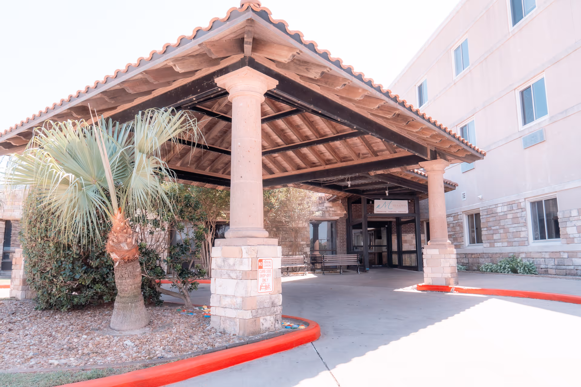 Covered entrance area of a senior living facility with stone pillars and a tiled roof. There is a palm tree and some bushes on the left side, a bench near the entrance, and a building with multiple windows in the background.