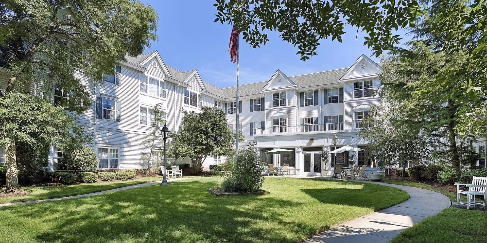 A bright and sunny courtyard area of a senior living facility with a well-maintained lawn, trees, and shrubs. The three-story white building with multiple windows surrounds the courtyard. There are benches and tables with umbrellas for outdoor seating, and an American flag is flying on a flagpole in the center.