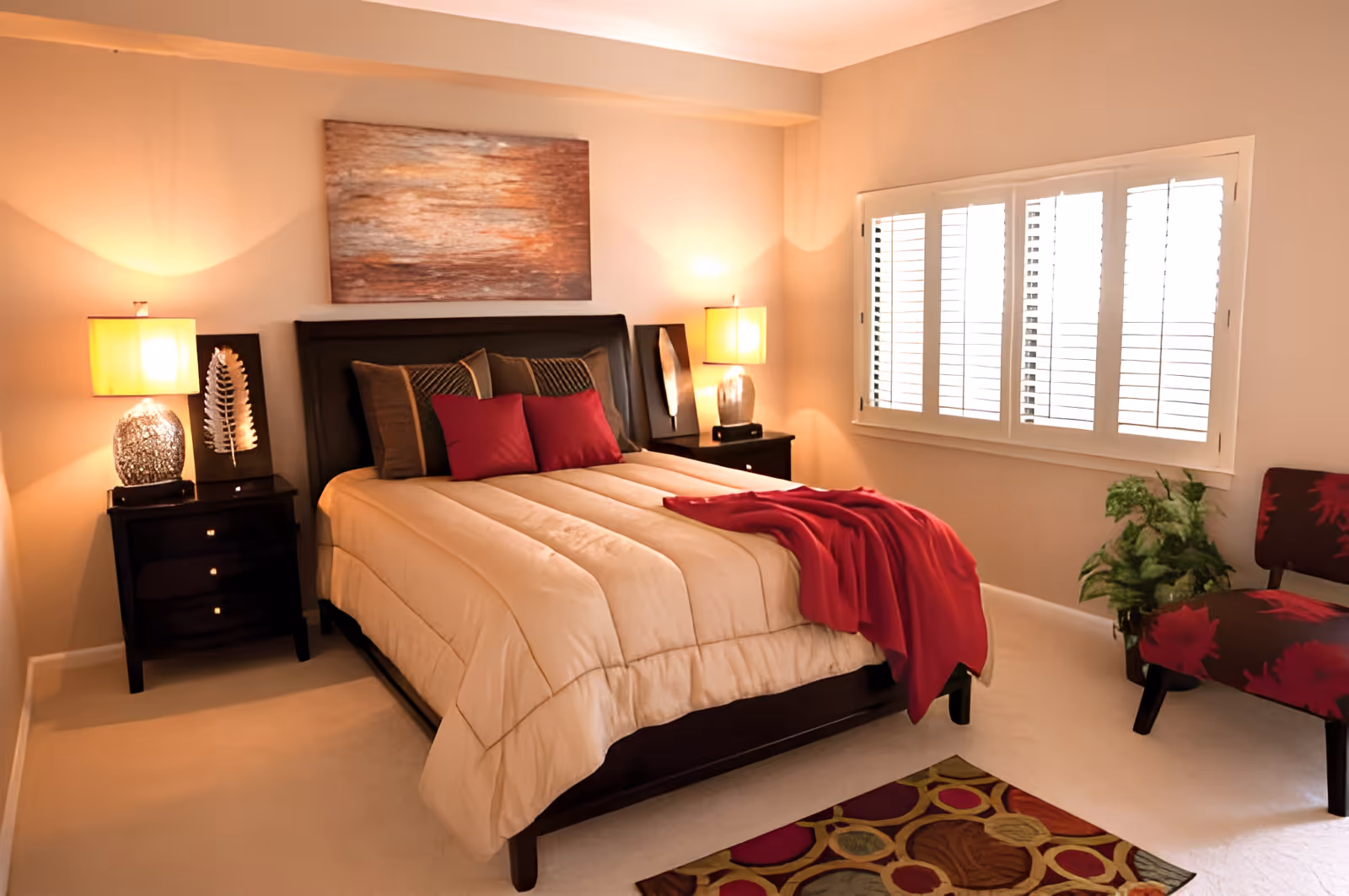 A cozy bedroom with a neatly made bed featuring beige bedding and red accent pillows and throw. Two dark wooden nightstands with lamps flank the bed. A large abstract painting hangs above the headboard. The room has beige walls, a window with white shutters, a patterned rug on the floor, a red and black floral chair, and a green potted plant in the corner.