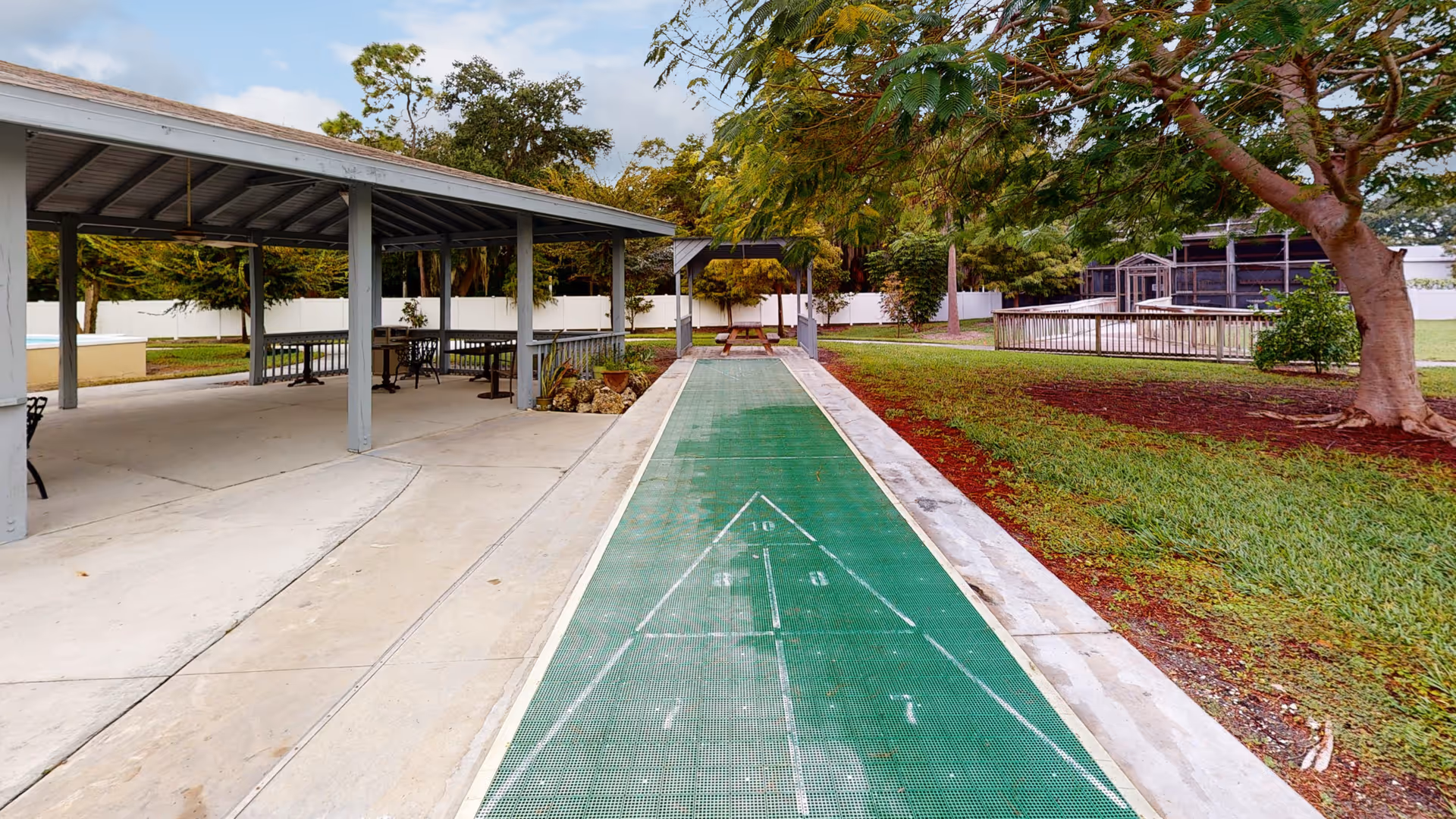 Outdoor recreational area featuring a covered pavilion with picnic tables on the left and a green shuffleboard court running alongside a grassy area with trees and a fenced enclosure in the background under a partly cloudy sky.