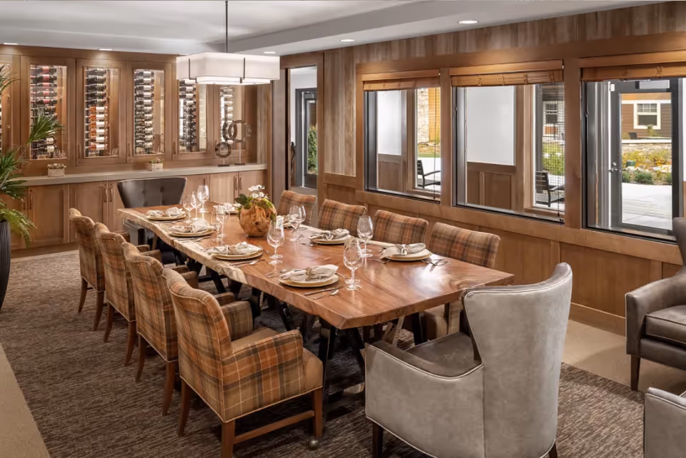 A spacious dining room with a large wooden table set for eight people. The table is surrounded by cushioned chairs, some with plaid upholstery and others in solid gray. The room features wood-paneled walls, large windows letting in natural light, and a built-in wine rack cabinet along one wall. A modern rectangular light fixture hangs above the table.
