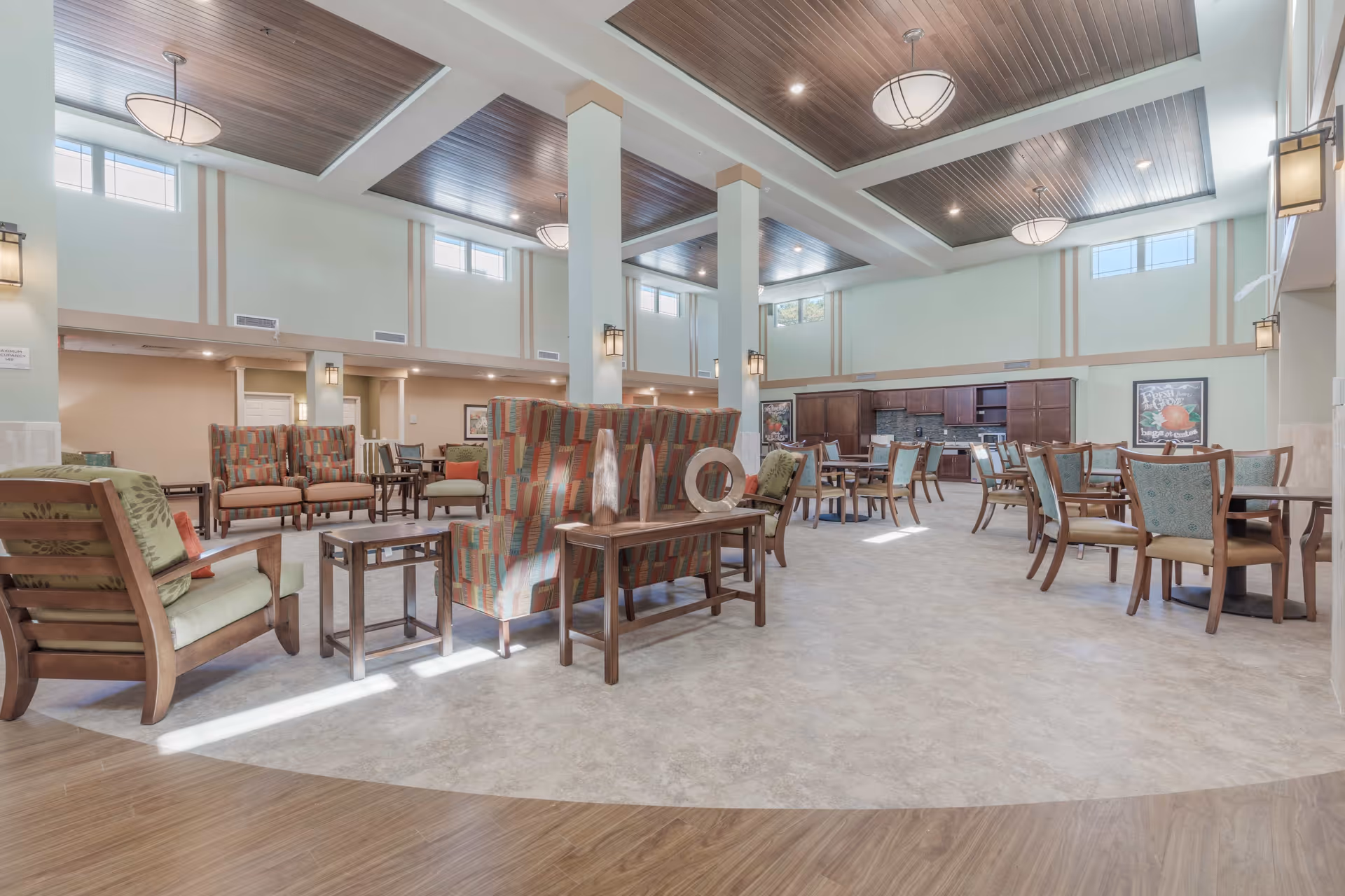A spacious and well-lit common area in a senior living facility featuring multiple seating arrangements with cushioned chairs and wooden tables. The room has high ceilings with wooden paneling and modern light fixtures. There are large windows near the ceiling allowing natural light to enter. In the background, there is a kitchenette area with dark wood cabinets and a countertop. The floor has a combination of wood and tile patterns.