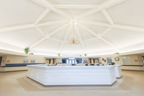A spacious, well-lit reception or common area with a large white octagonal desk in the center under a high, white vaulted ceiling with a chandelier. The room has beige walls, carpeted floors, and some plants and framed pictures on the walls.