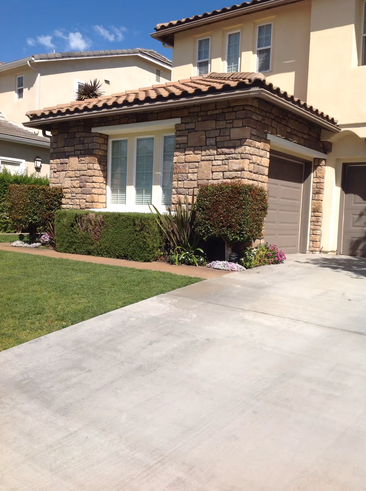 Exterior view of a residential building with a stone facade, beige walls, and a tiled roof. There are two garage doors, a window with white shutters, trimmed bushes, and a well-maintained lawn under a blue sky with some clouds.