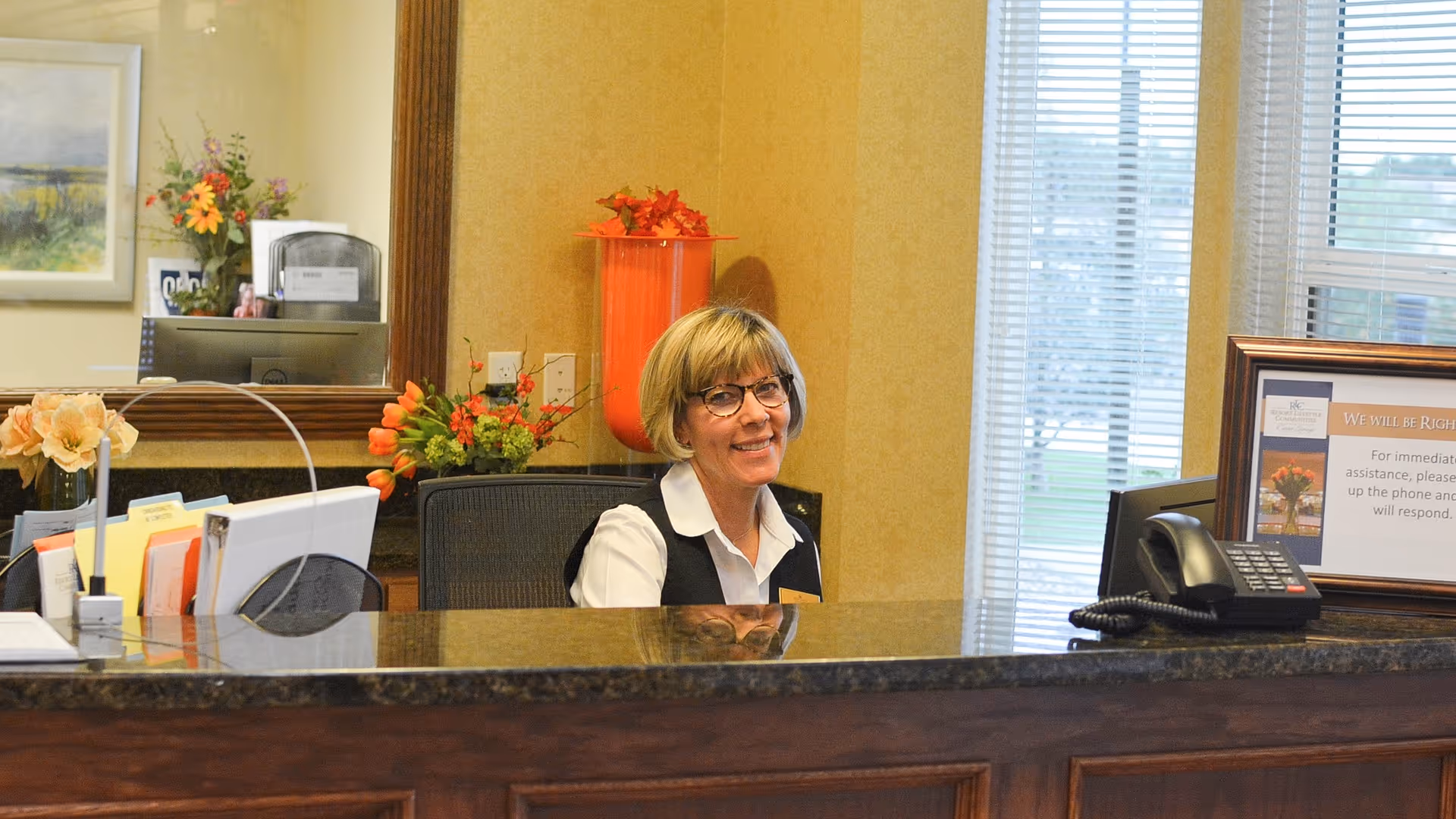 A smiling woman with short blonde hair and glasses sits behind a reception desk in a well-lit room with beige walls. The desk has a dark granite countertop with organized folders, a telephone, and a framed sign. Behind her, there are flowers, a large mirror, and a window with blinds letting in natural light.