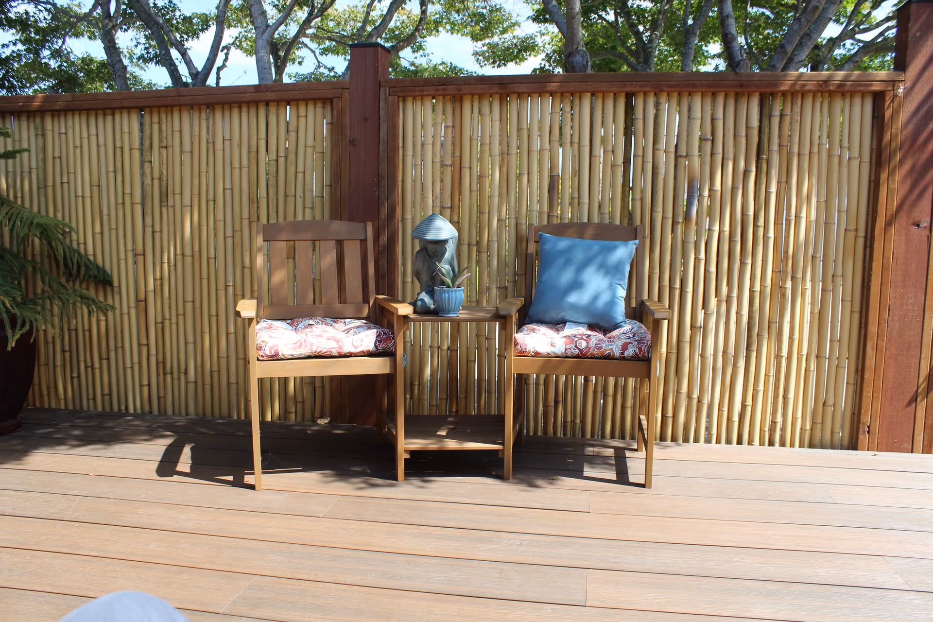 Two wooden chairs with patterned seat cushions and a blue pillow on one chair, placed on a wooden deck with a small wooden table between them holding a decorative item and a small potted plant. The background features a bamboo fence with wooden posts and trees visible above the fence.