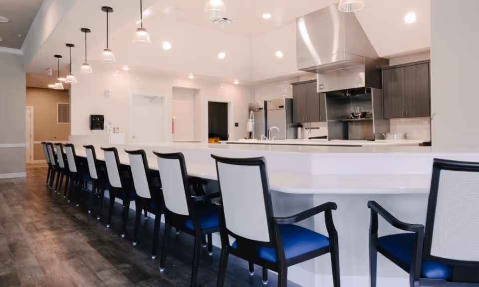 A modern kitchen area with a long white counter and multiple high-back chairs with blue seats lined up along it. The kitchen features stainless steel appliances, including a large stove hood, and dark wood cabinets. Pendant lights hang from the ceiling above the counter, and the floor has a dark wood finish.