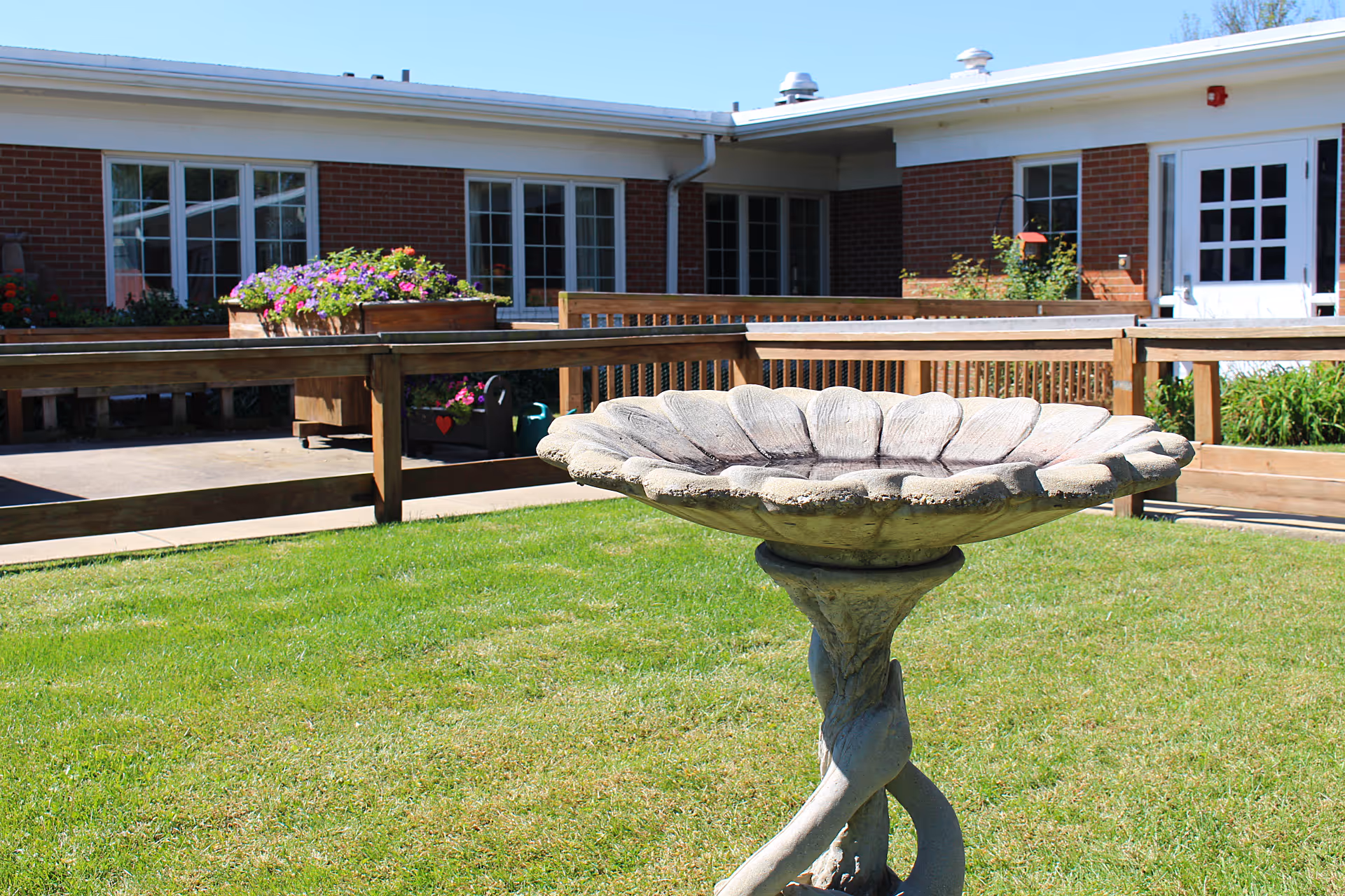 A birdbath shaped like a large flower on a green lawn in front of a single-story brick building with white-framed windows and a wooden railing. There are flower boxes with colorful flowers near the building.