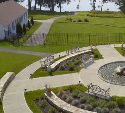 Outdoor garden area with curved concrete pathways, multiple wooden benches, landscaped plants, a circular stone water feature, and a view of a lake with boats in the background.