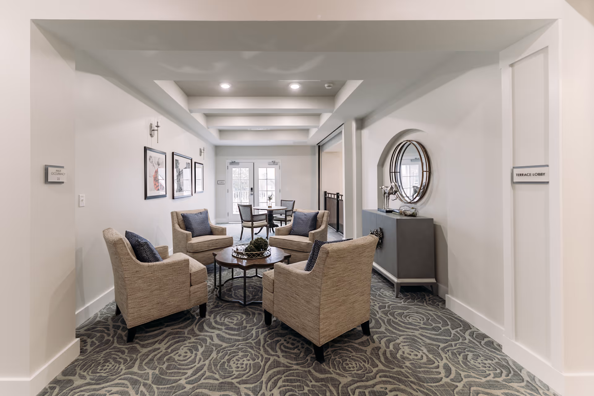 A cozy indoor seating area with four beige armchairs arranged around a round wooden coffee table on a patterned carpet. The walls are white with framed artwork on one side and a gray console table with a round mirror and decorative items on the other. In the background, there is a small dining table with four chairs near double glass doors leading outside.