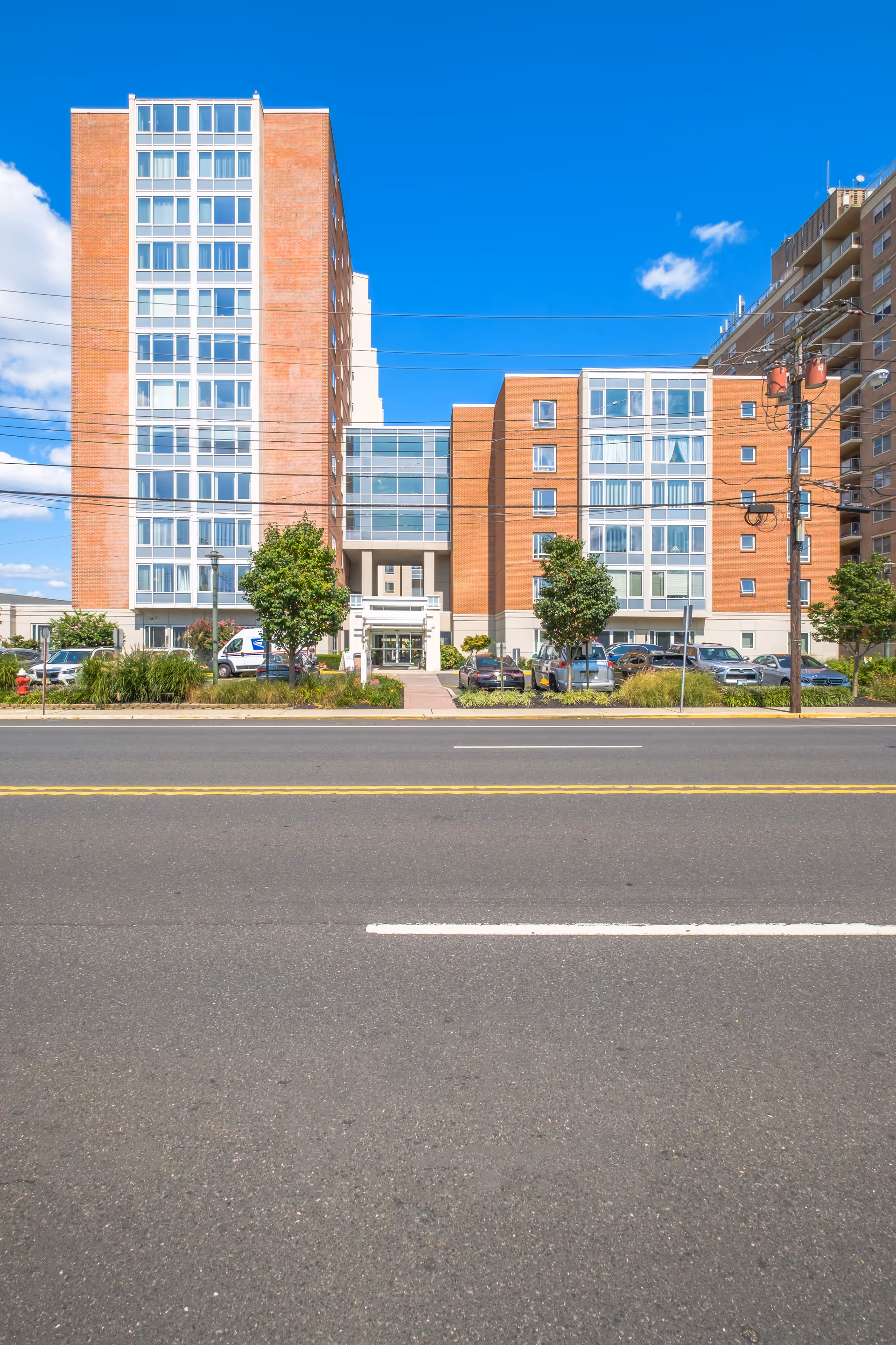 Front exterior of a multi-story brick senior living building with a central glass atrium and parked cars under a clear blue sky.