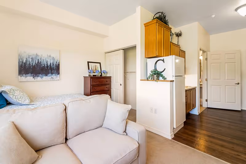 Interior view of a senior living facility room at The Chesterley showing a beige couch in the foreground, a bed with pillows and a forest-themed painting on the wall, a wooden dresser with decorative items, a closet with sliding doors, and a small kitchen area with a refrigerator and wooden cabinets.