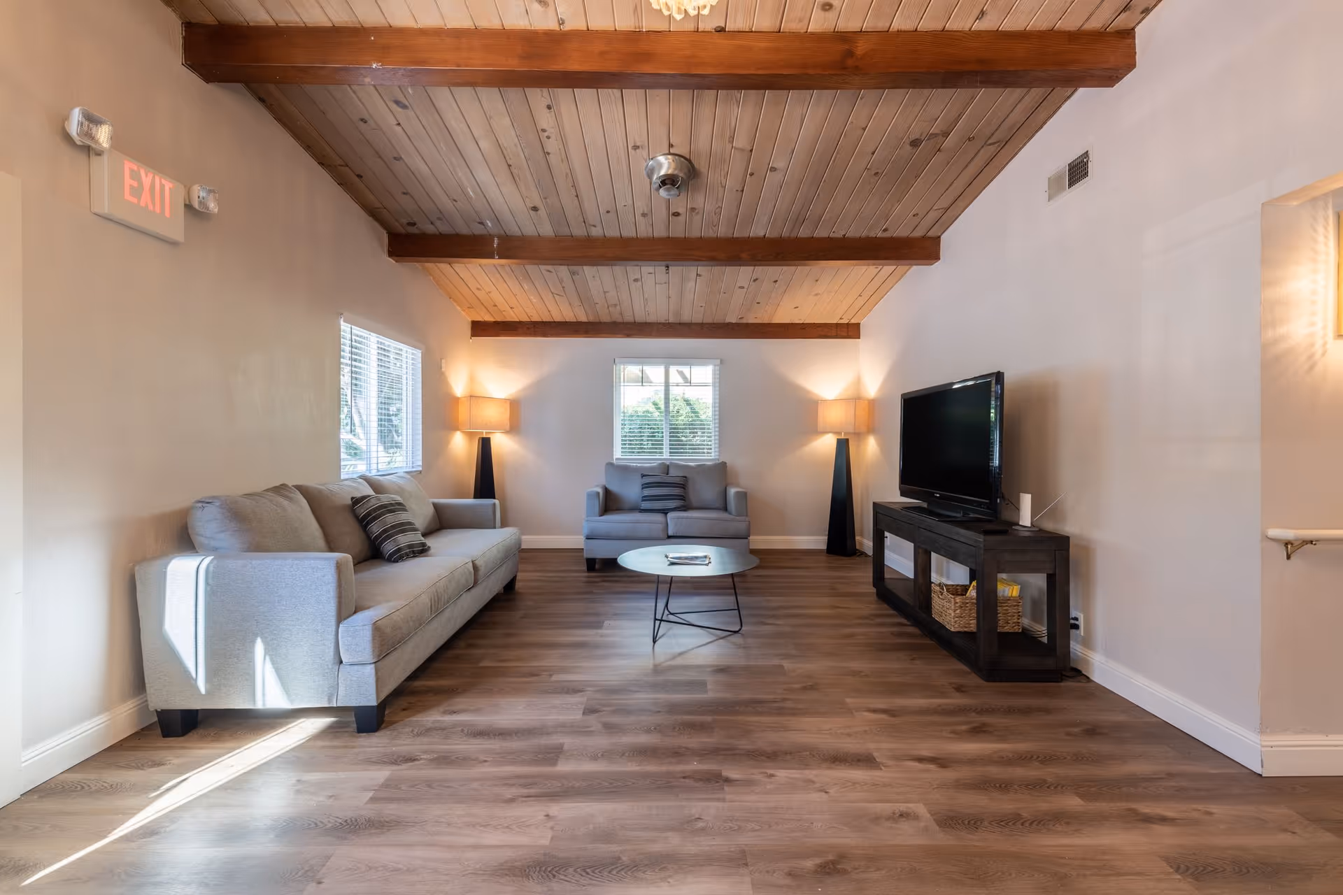 A cozy living room with wooden ceiling beams, two gray sofas with striped pillows, a round coffee table in the center, two floor lamps with beige shades, a flat-screen TV on a dark wooden TV stand with baskets underneath, and windows letting in natural light.