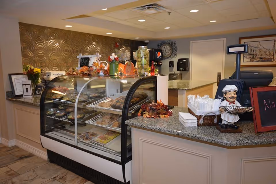 A bakery display case filled with various baked goods in a retirement community setting. The counter has decorative autumn leaves, a small chef figurine, napkins, and a cash register. The background features a textured wall, a clock, and a soda dispenser.