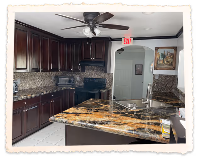 A modern kitchen with dark wooden cabinets, a granite countertop island with a built-in sink, a ceiling fan with lights, a microwave, and an electric stove. There is an exit sign above an arched doorway leading to another room, and a framed painting on the wall.