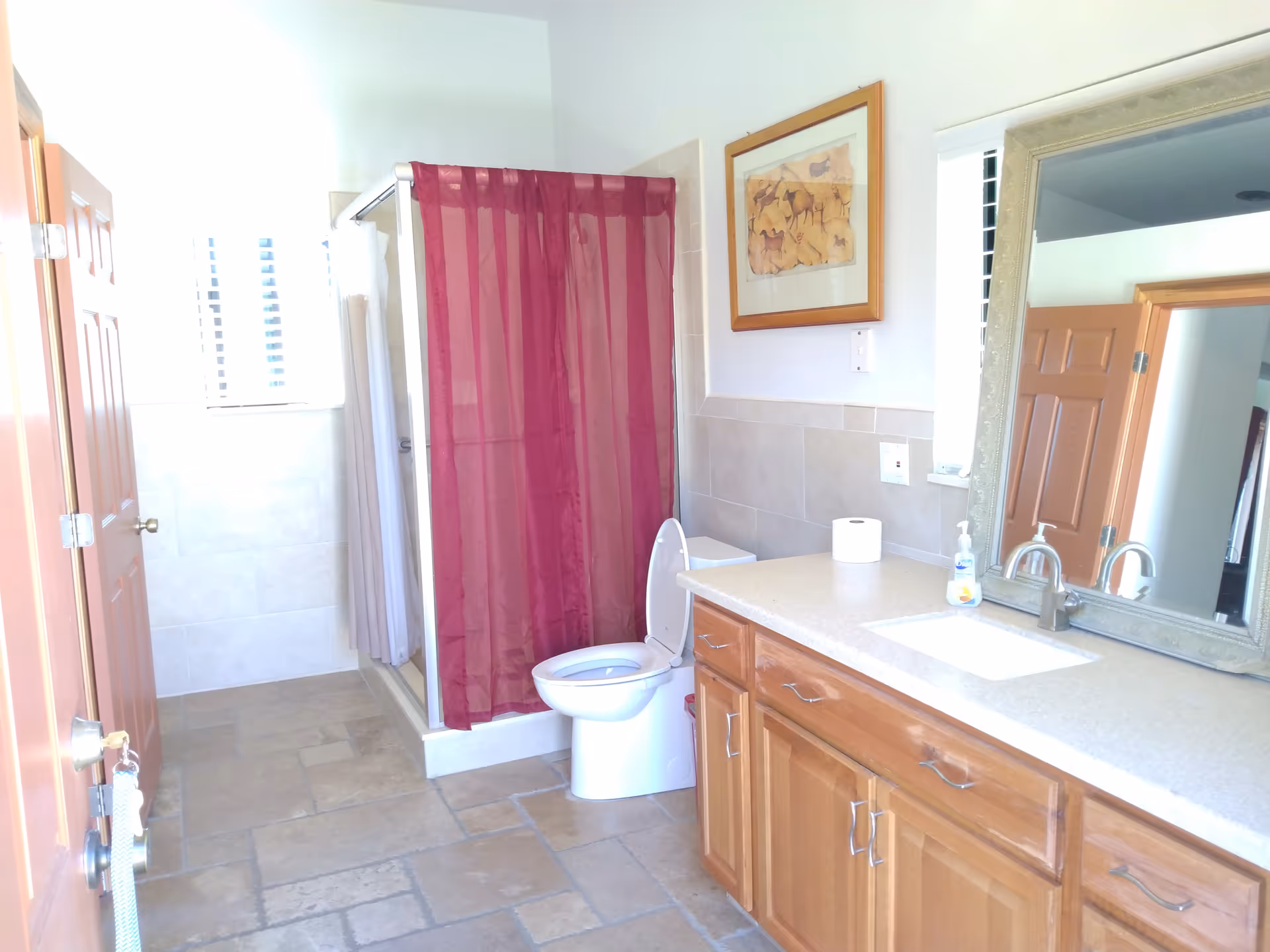 A bright bathroom with a tiled floor, wooden cabinets with a countertop and sink, a large mirror above the sink, a toilet with the lid open, and a shower with a red curtain. There is a framed picture on the wall and a window letting in natural light.