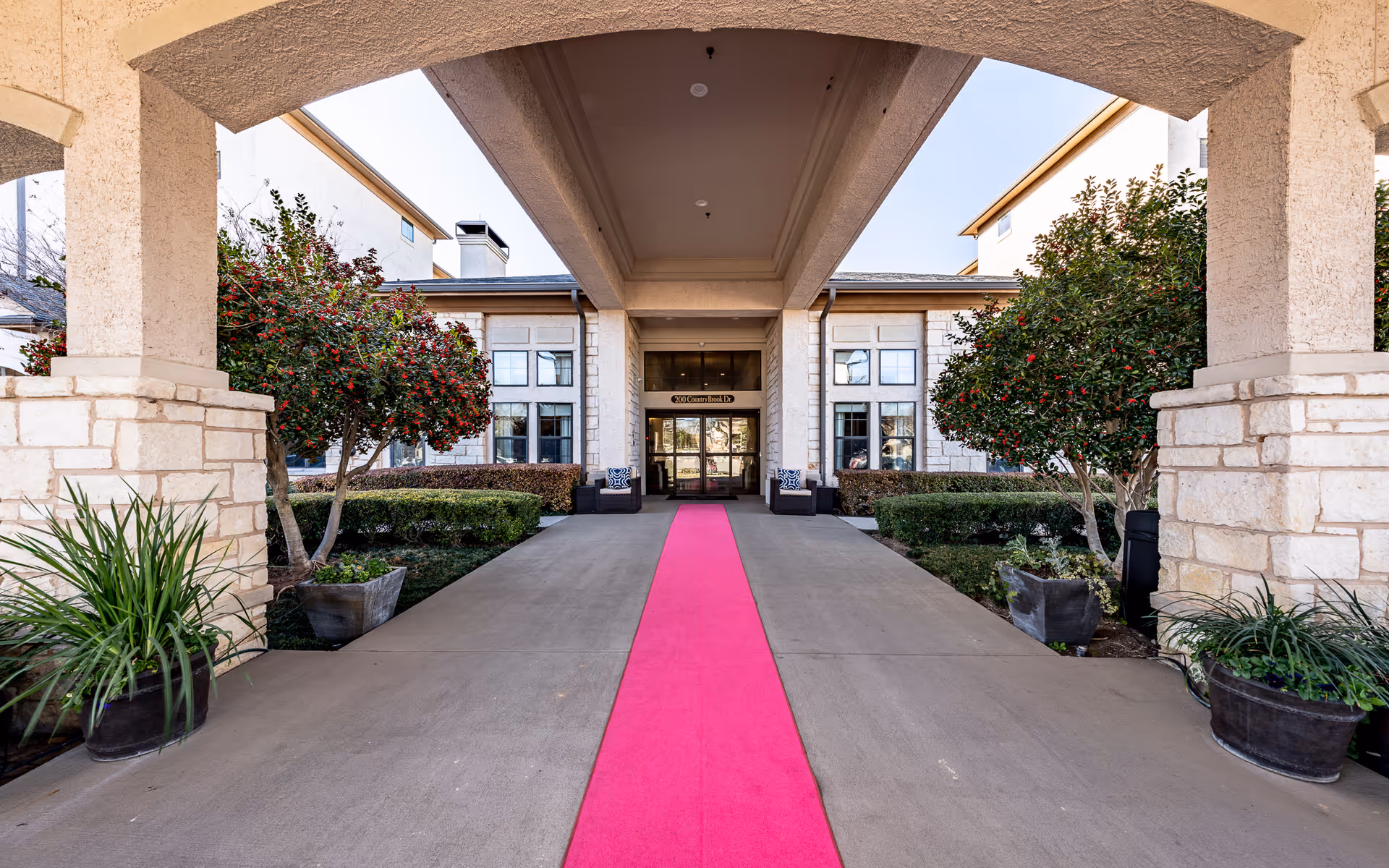 Entrance of Conservatory At Keller Town Center with a covered driveway featuring a bright pink carpet runner, flanked by stone pillars and landscaped bushes and trees on both sides.